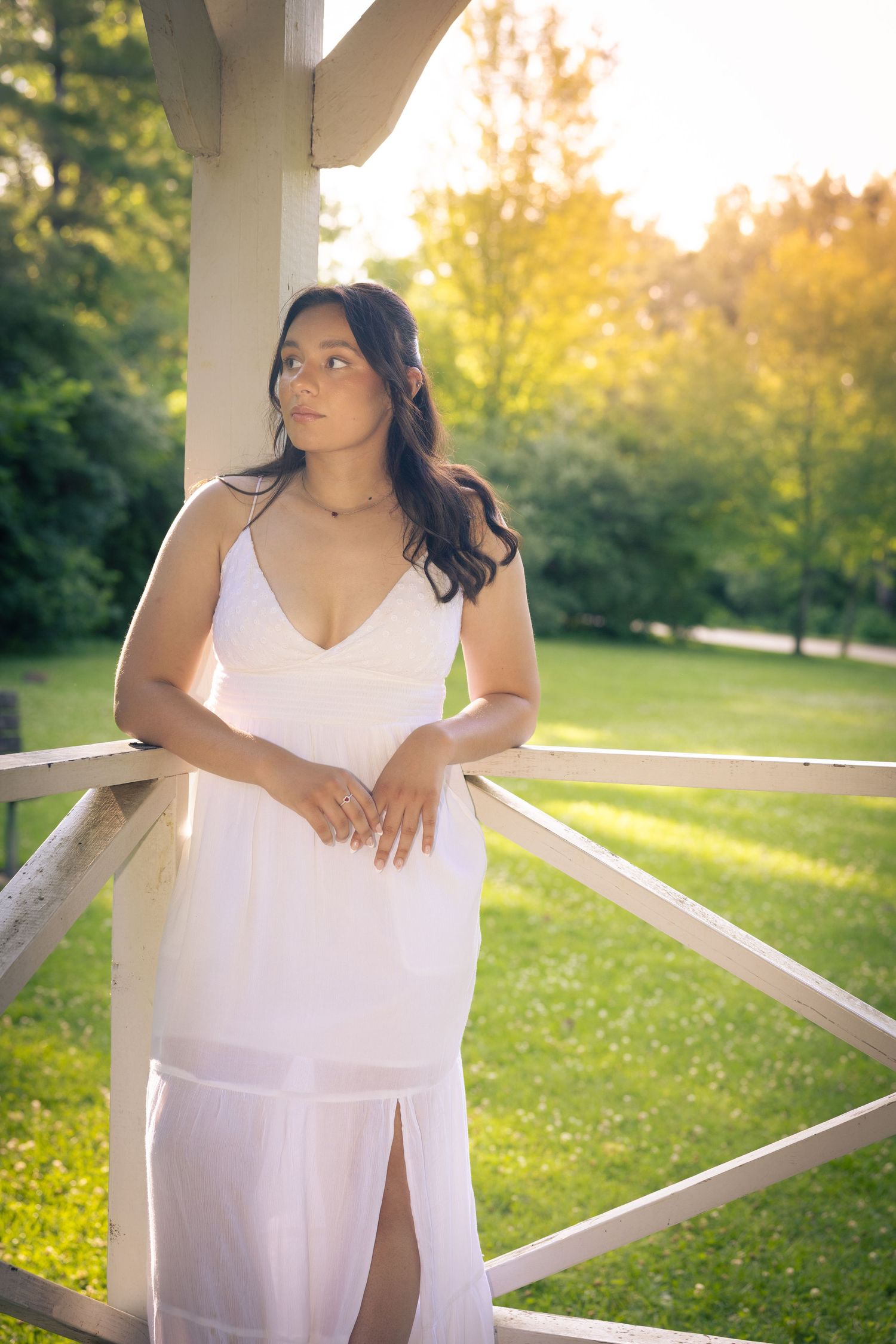 Young woman in white spaghetti strap dress poses gracefully by wooden railing in sunlit green park.