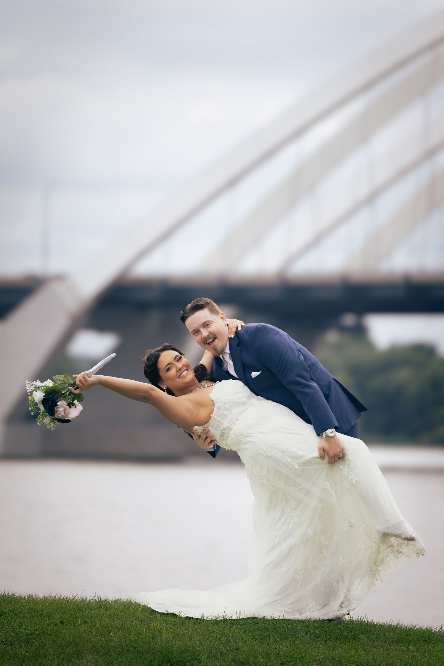 Groom dips bride in white gown outdoors near a bridge, both laughing joyfully on their wedding day.