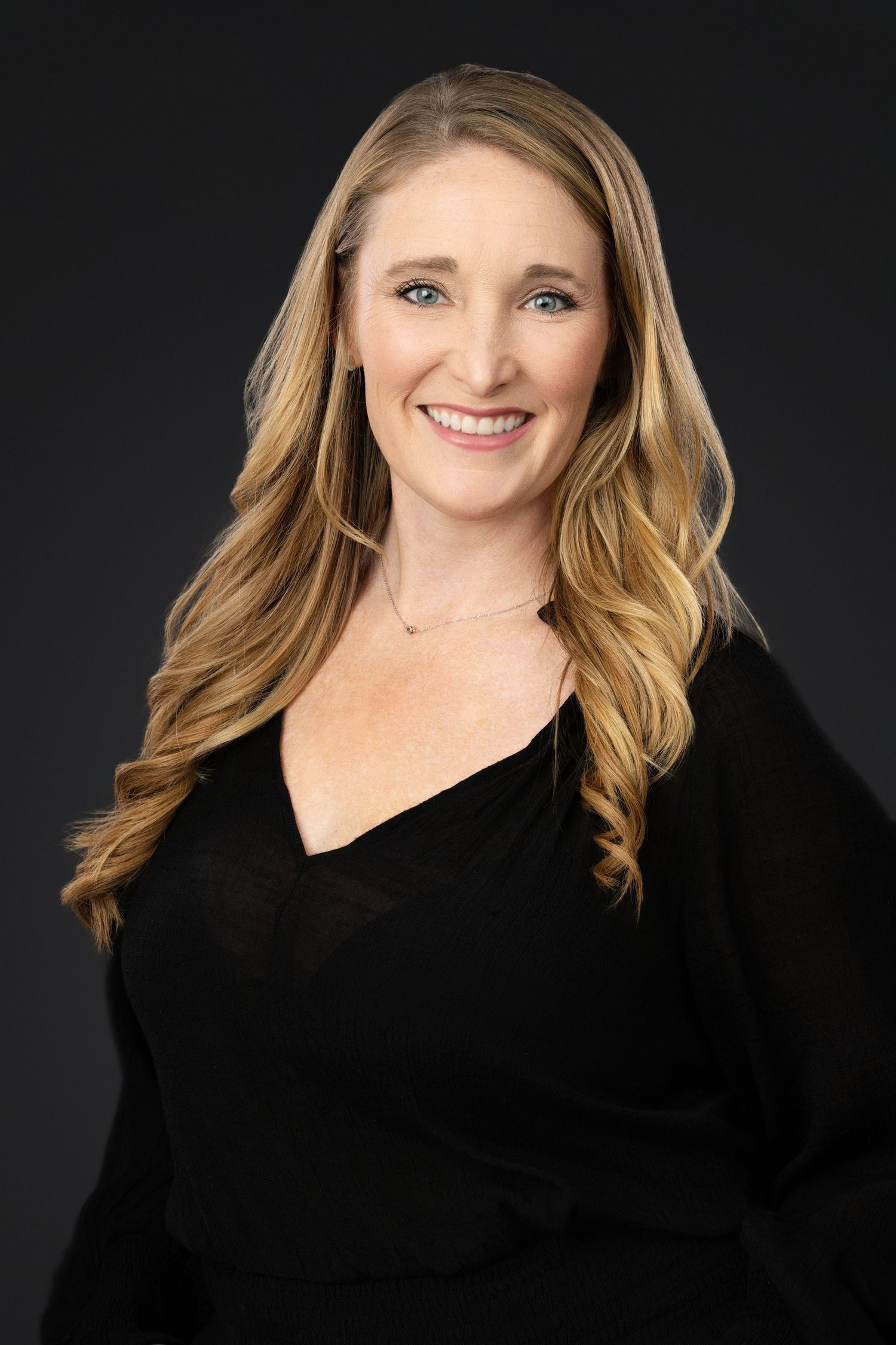 Professional studio portrait of woman with blonde wavy hair in black v-neck top against dark backdrop.