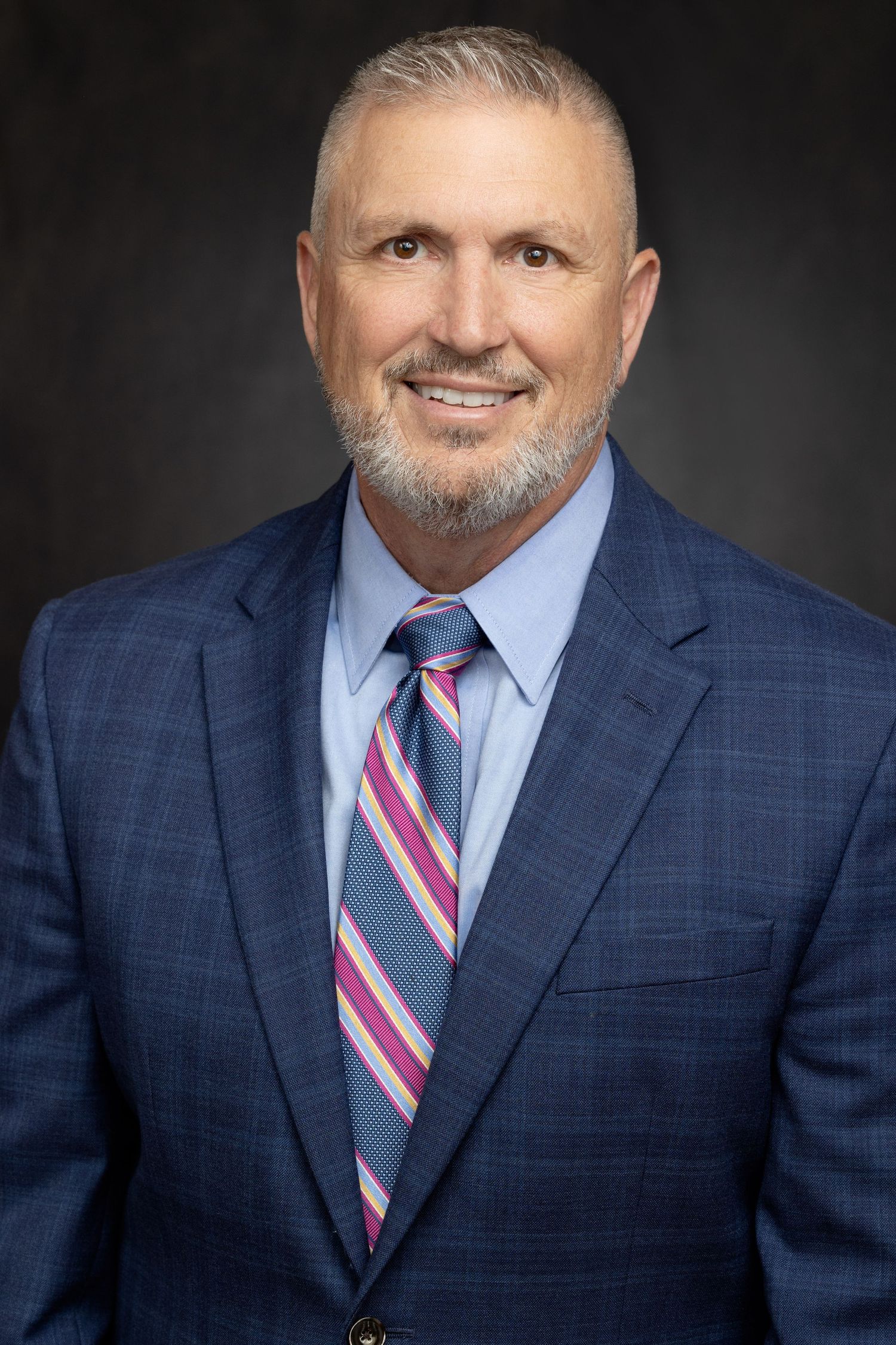 Professional headshot portrait of someone in blue plaid suit with striped pink tie against dark background.