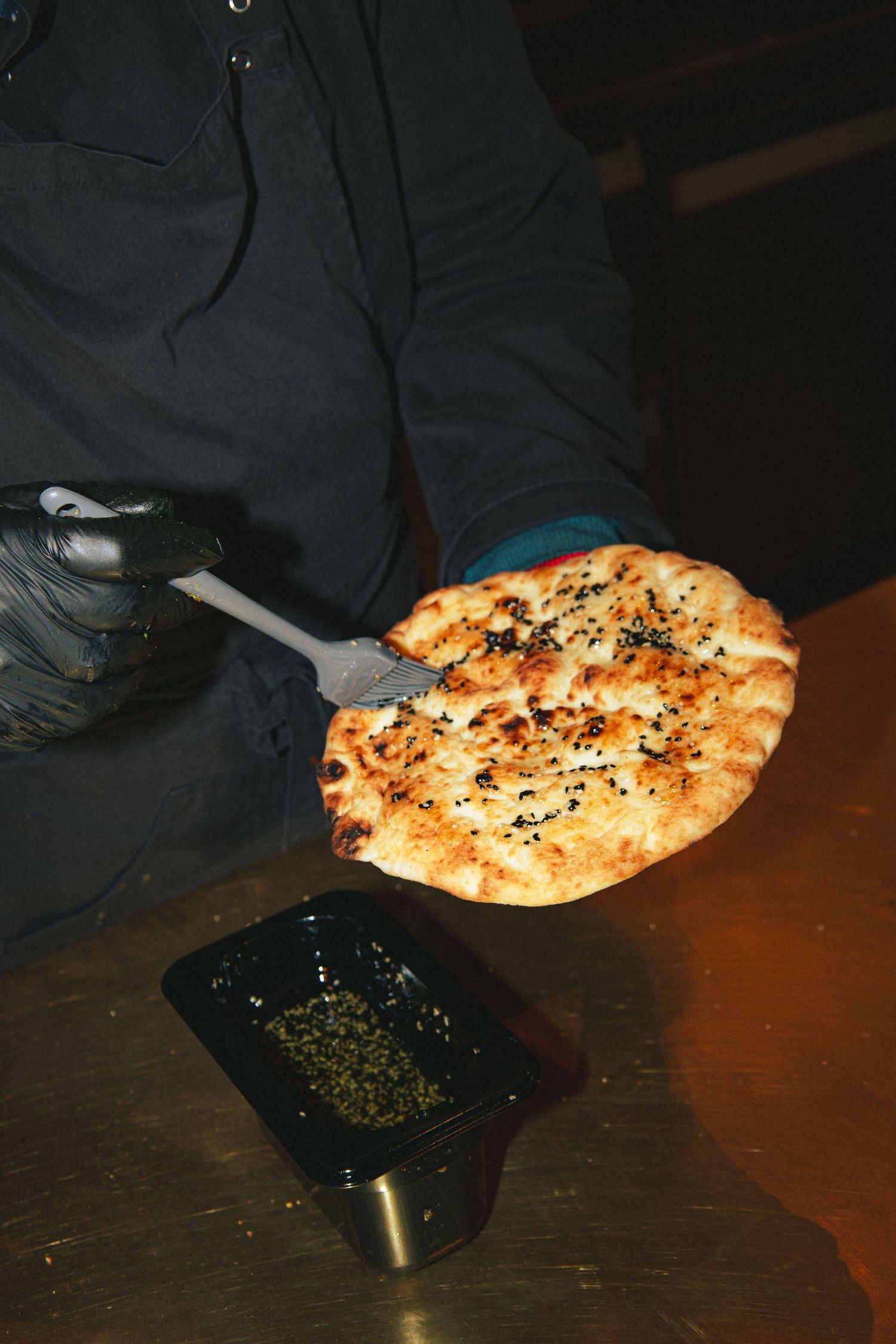 A flatbread topped with herbs and seasonings served alongside a small dish of spices on a dark table.