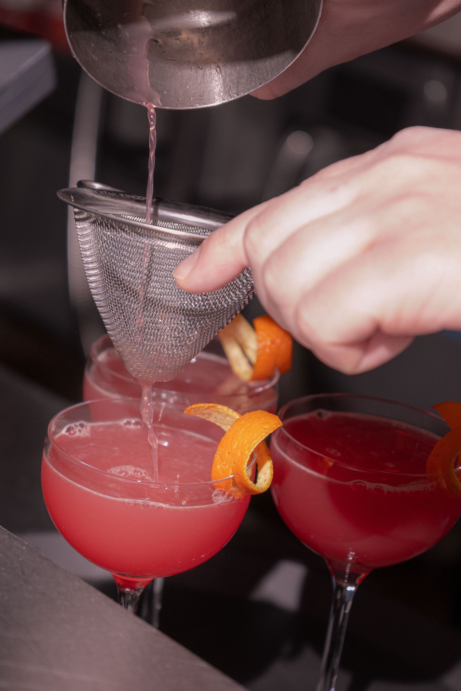 Bartender straining a pink cocktail through a mesh strainer into coupe glasses garnished with orange peel.