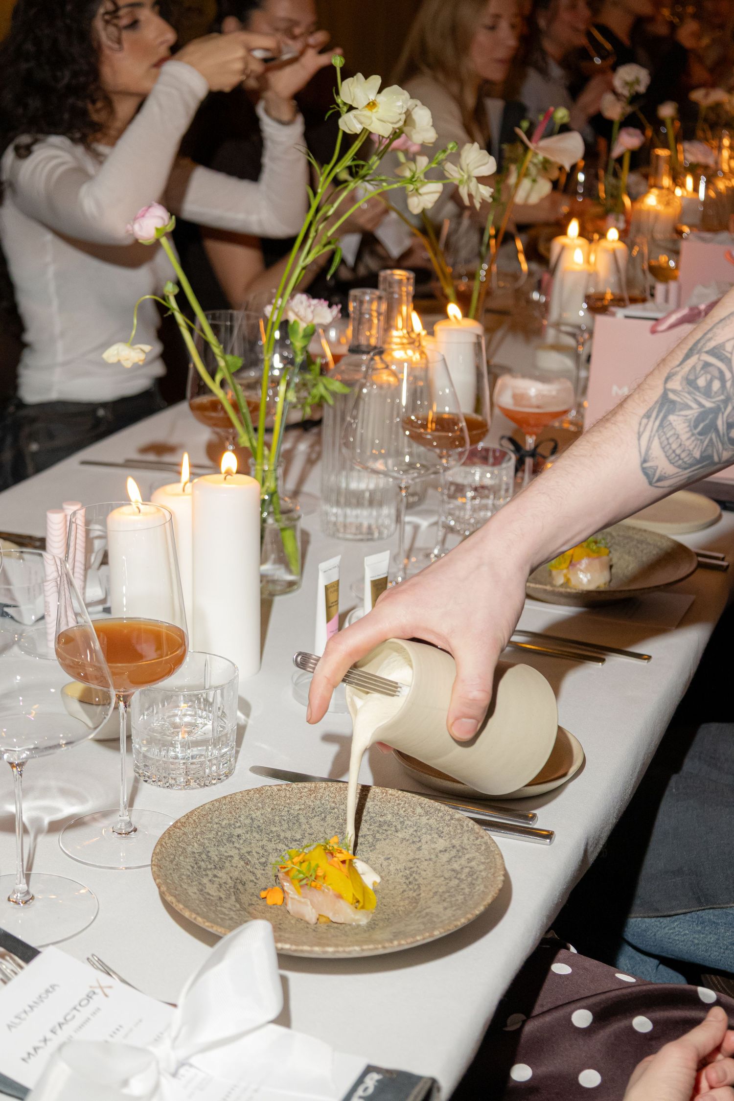 Elegant dinner party with candles, floral arrangements, and a server pouring sauce onto a gourmet dish at a long table.