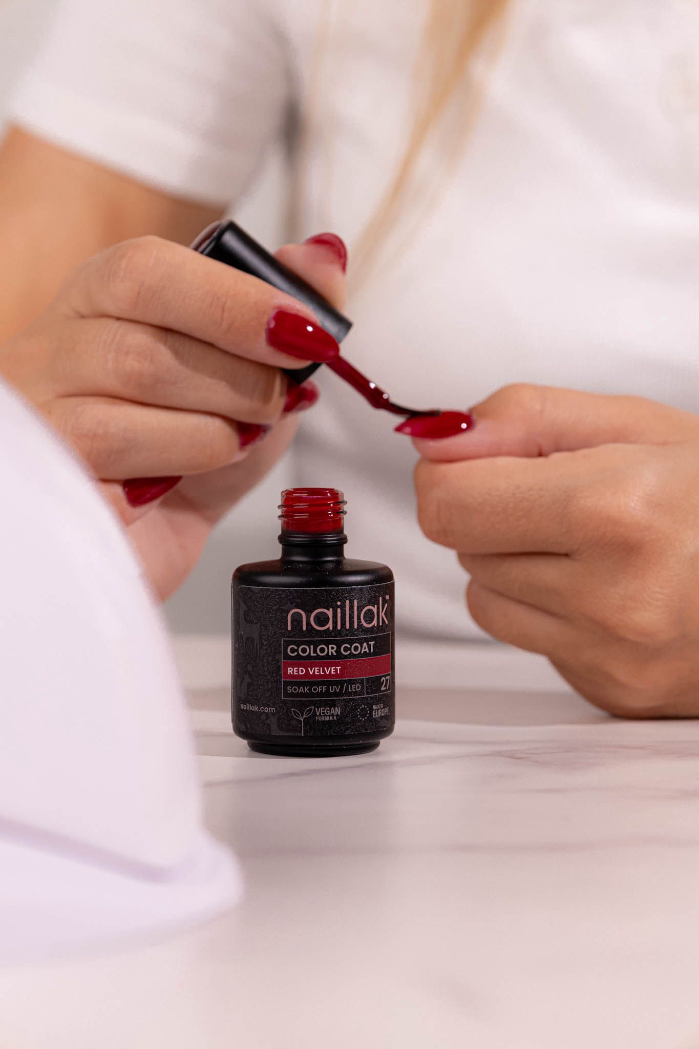 Hands opening a red nail polish bottle on white surface.