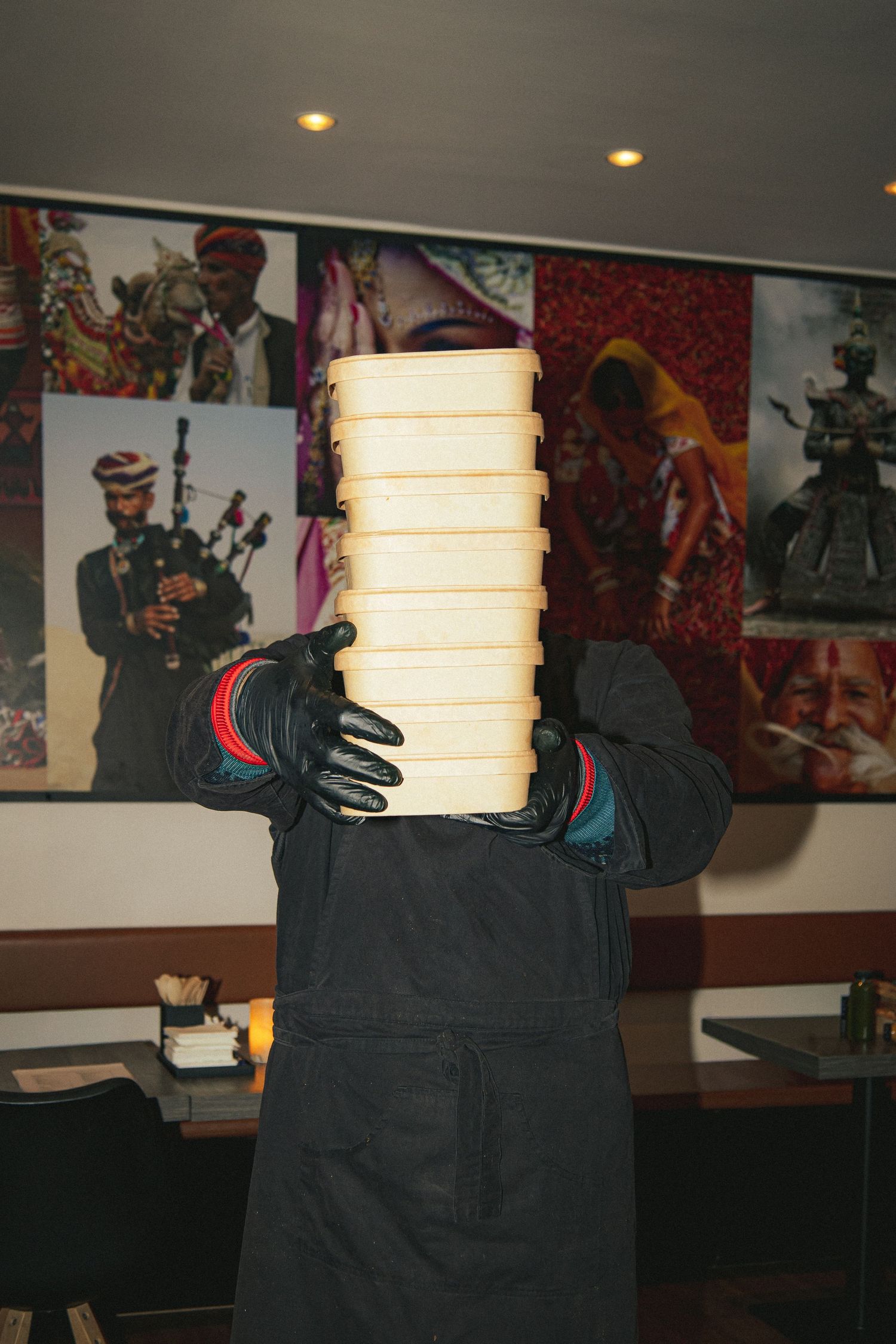 Someone in dark clothing and gloves holding a tall stack of white takeout containers in a restaurant.