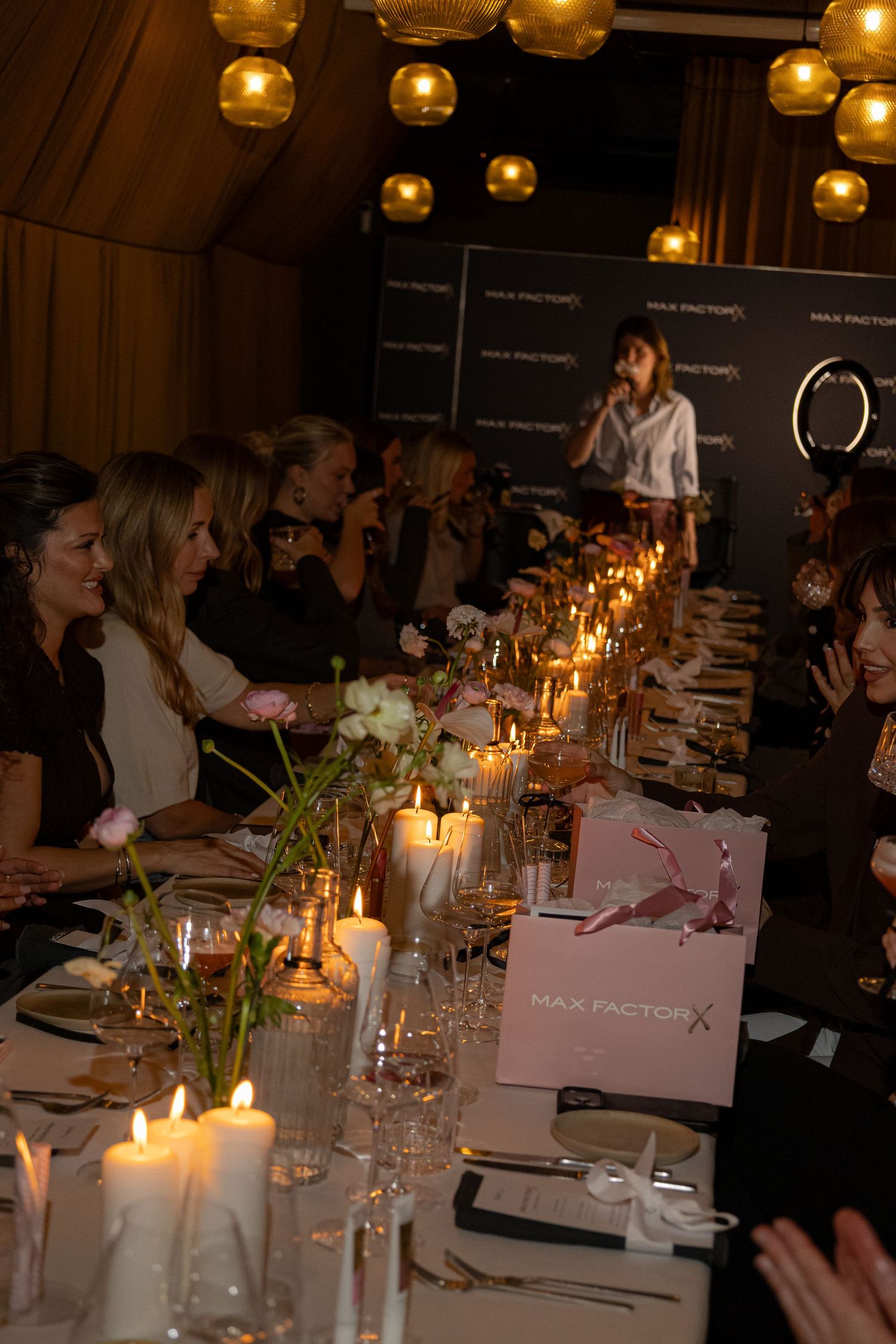 Elegant Max Factor dinner event with guests seated at a candlelit table, floral centerpieces, and a speaker at the end.