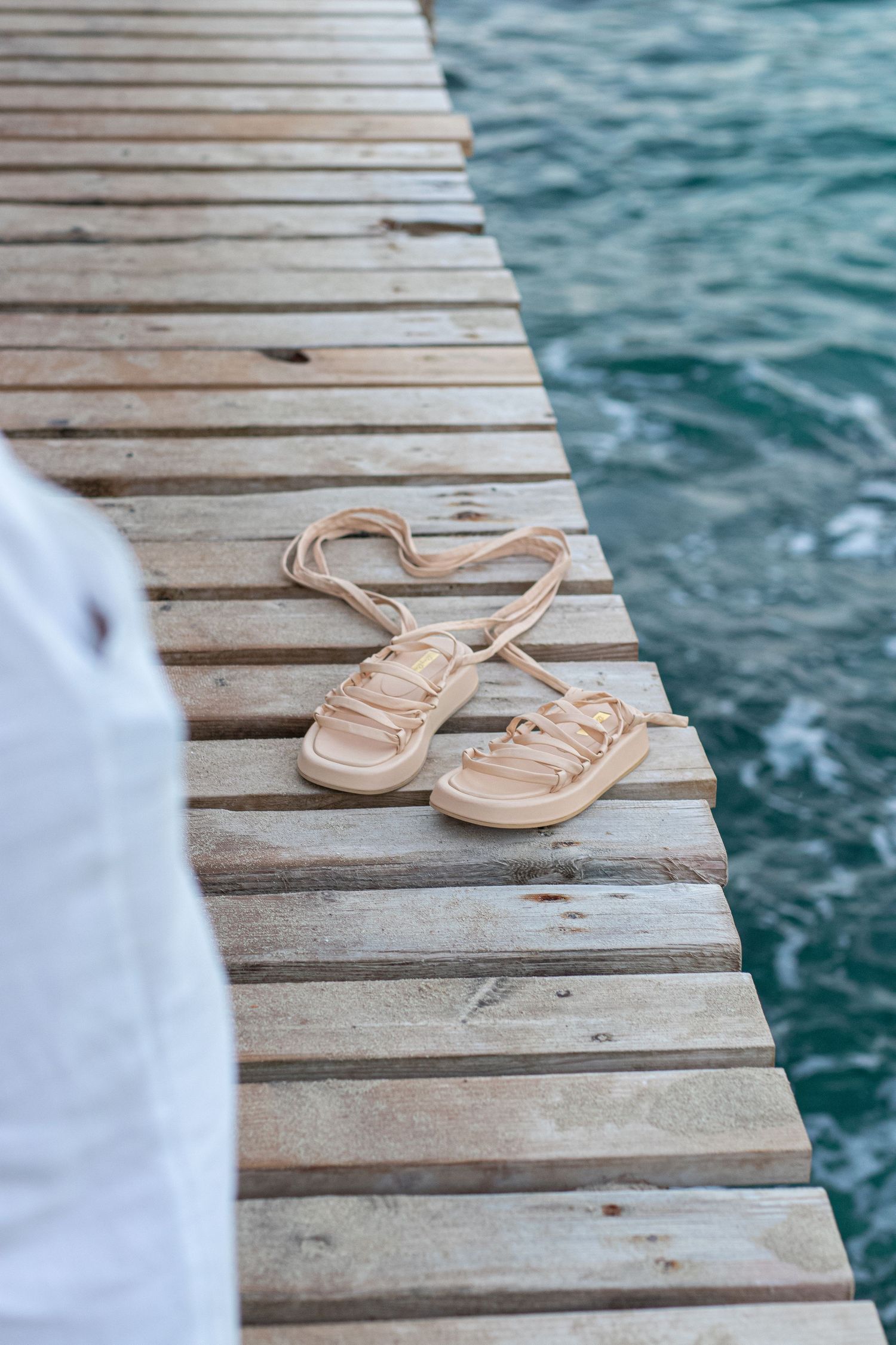 Beige sandals placed on wooden dock with blue water in background.