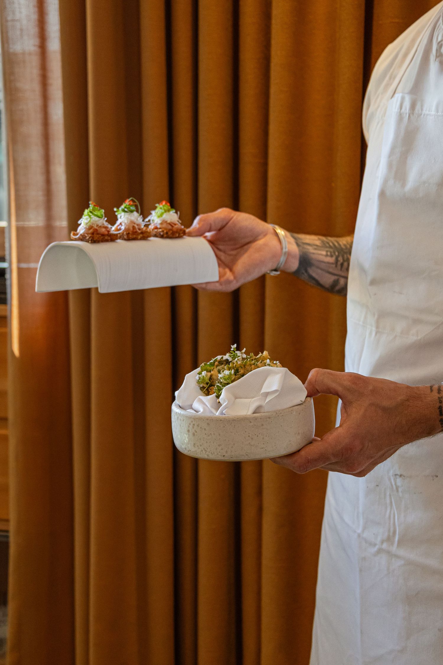 Tattooed chef in white uniform presenting elegant plated dishes with microgreens in modern restaurant.