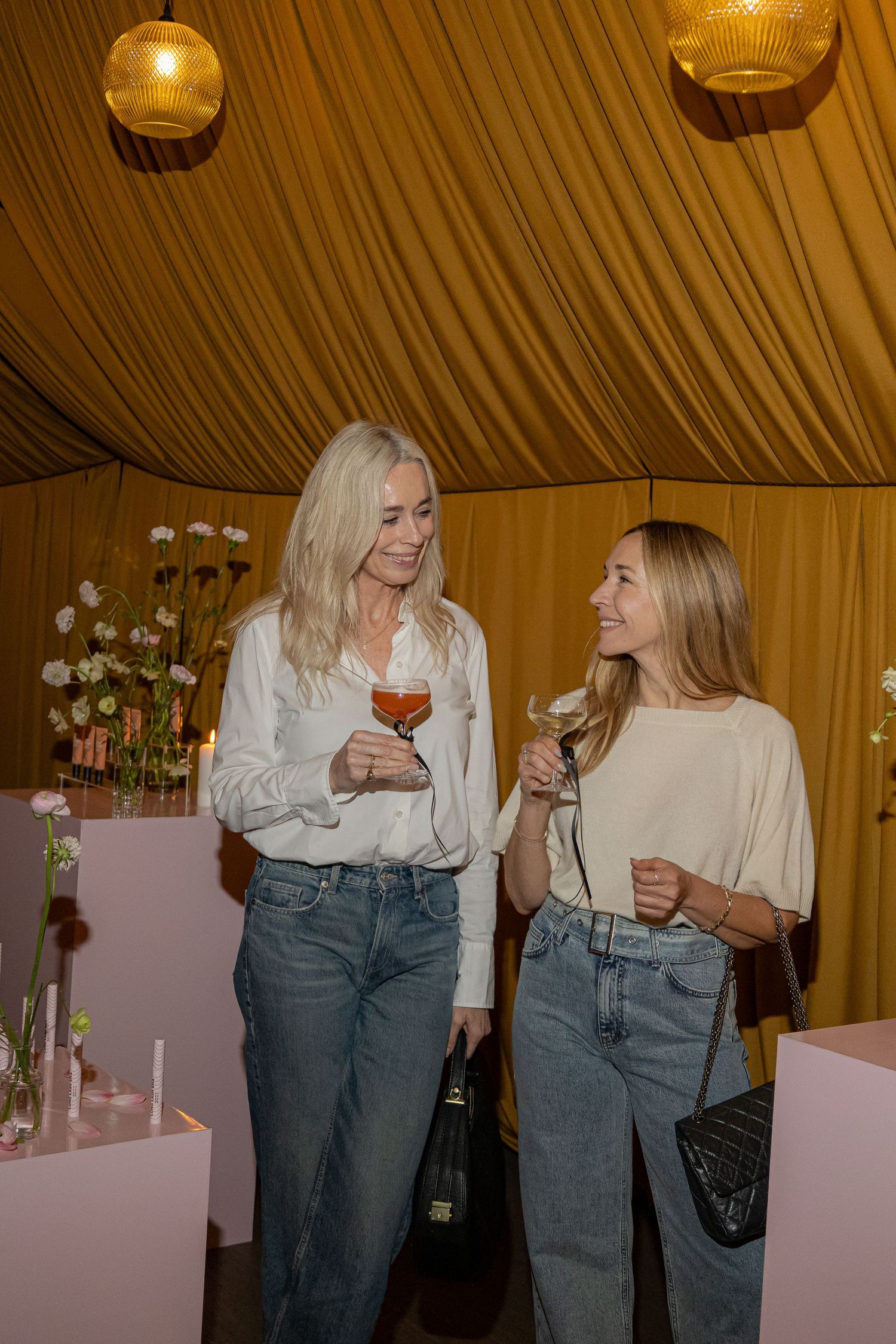 Two women holding wine glasses chat at an elegant event under a golden draped ceiling with floral decor.