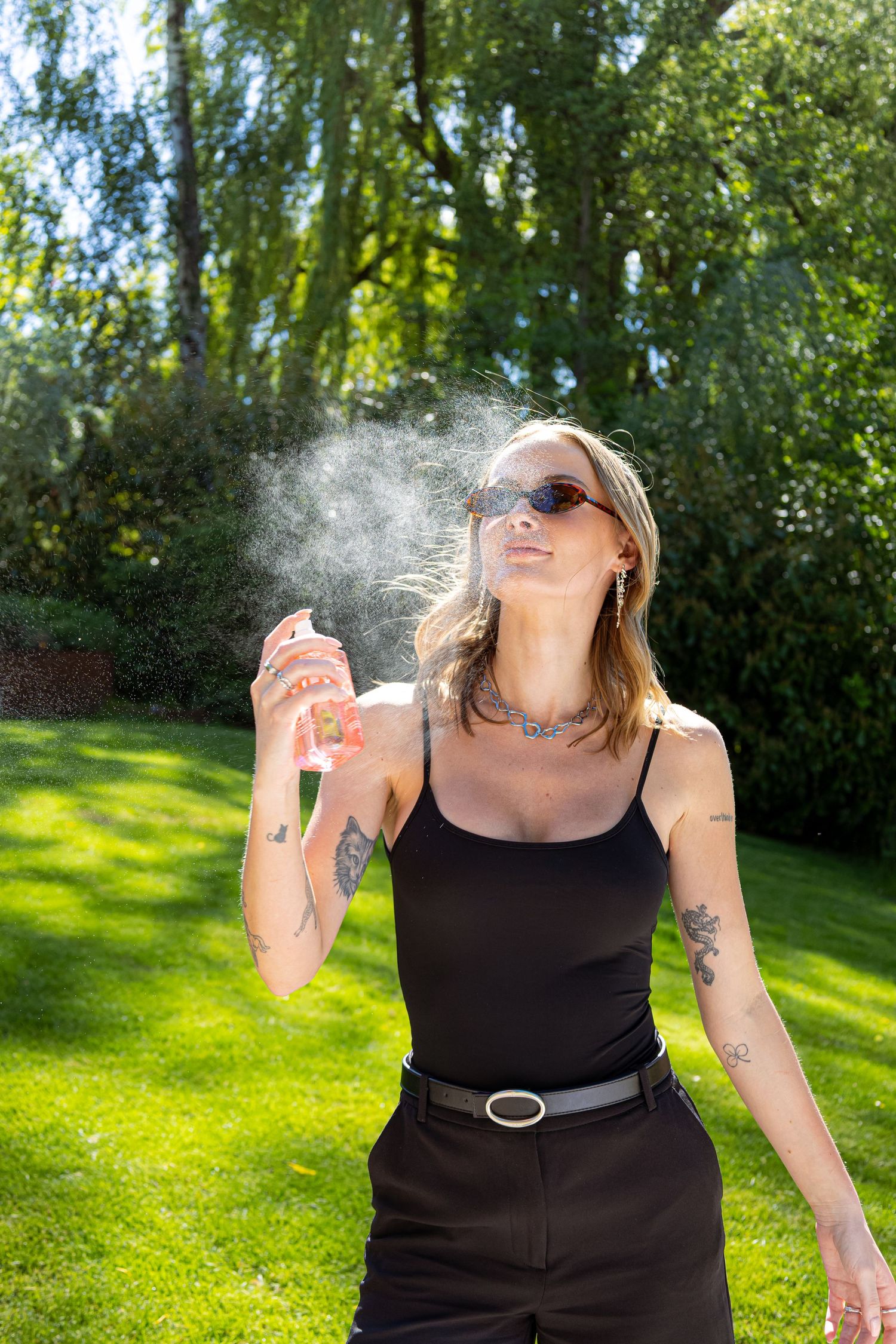 Tattooed woman in black tank top spraying mist in sunny garden, wearing sunglasses and layered necklaces.