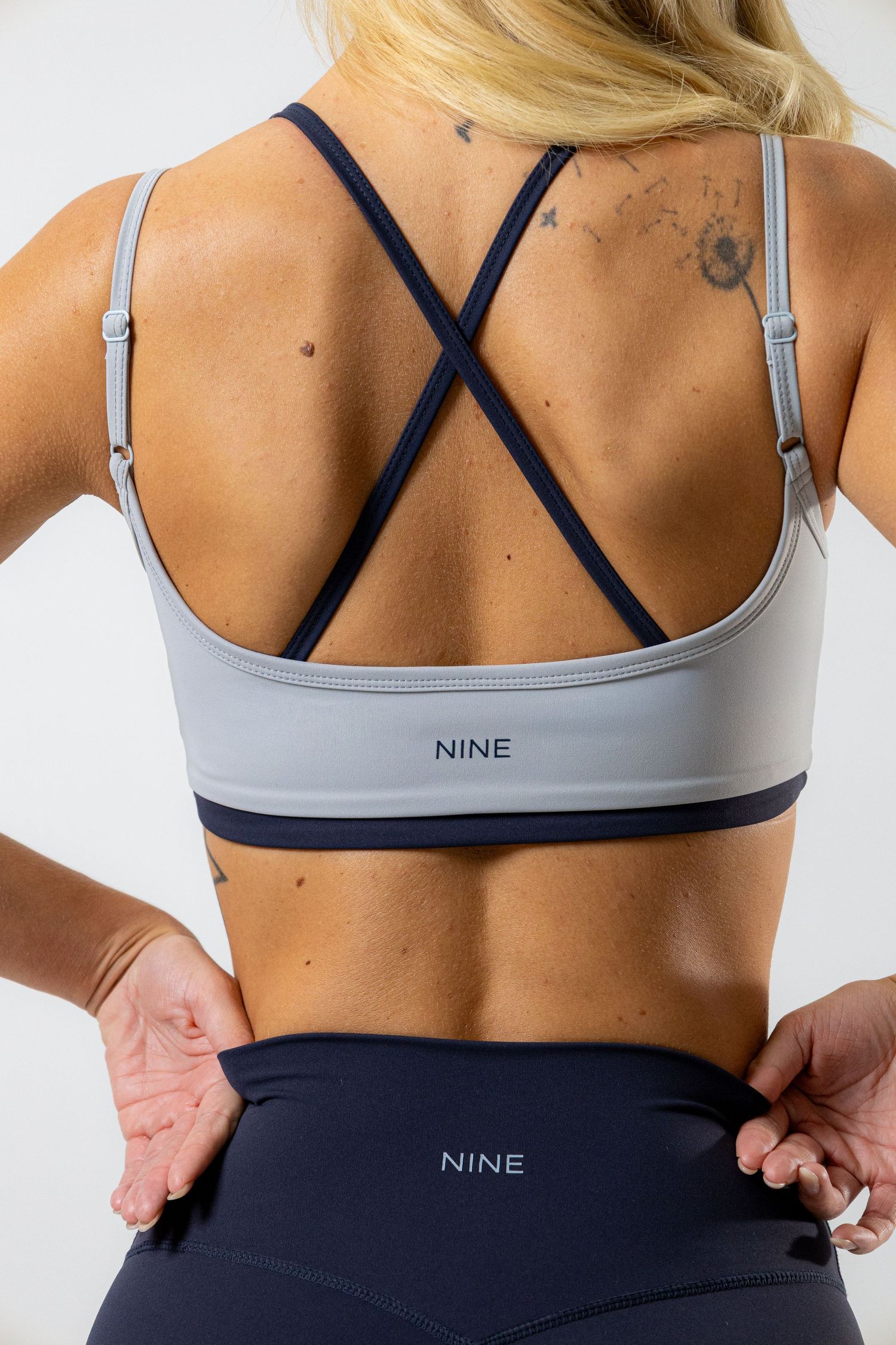 Woman wearing a light blue NINE sports bra with cross-back straps and navy blue NINE leggings, back view.