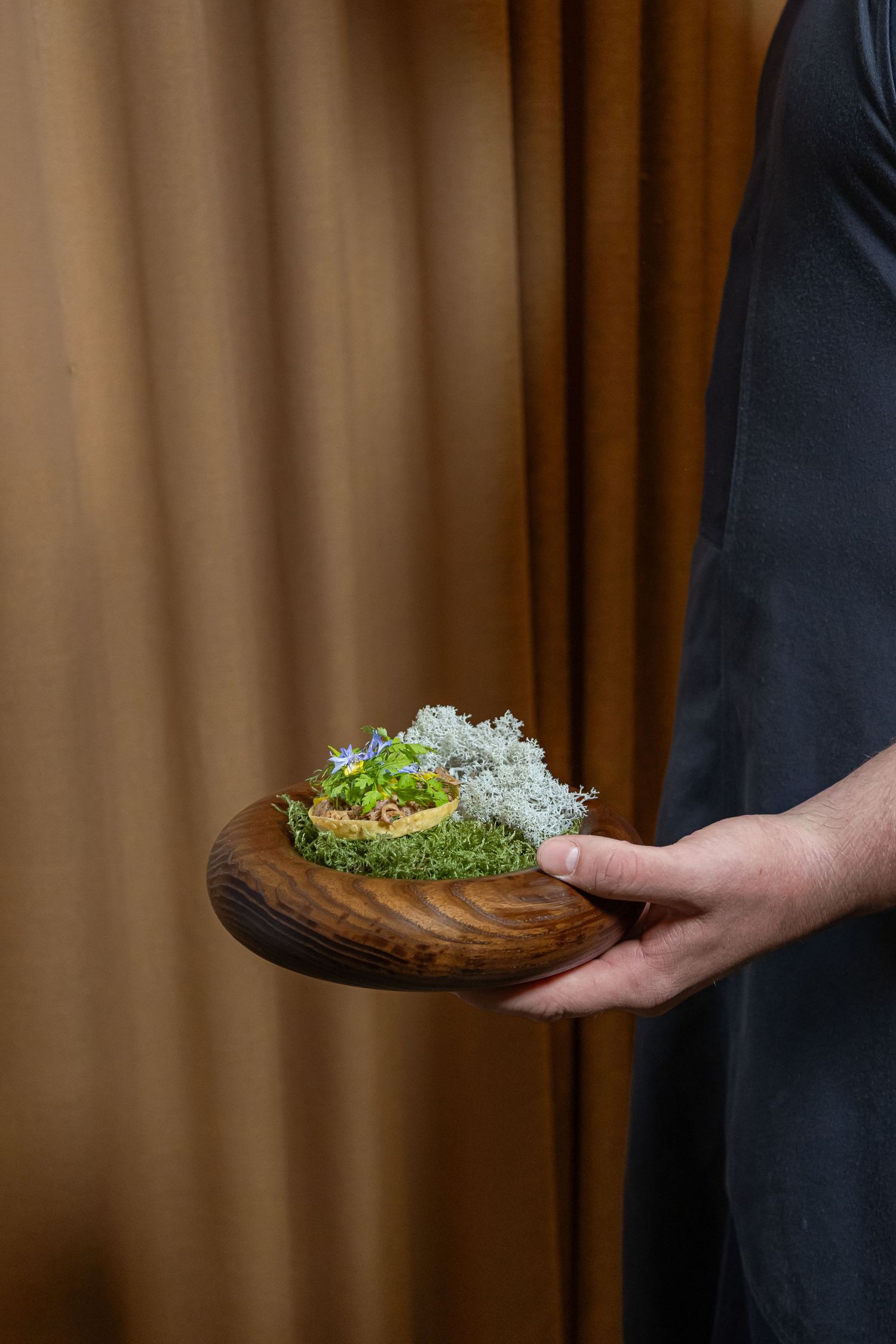 Chef holding a rustic wooden plate with an elegant micro-herb garnished dish, set against warm brown curtain backdrop.