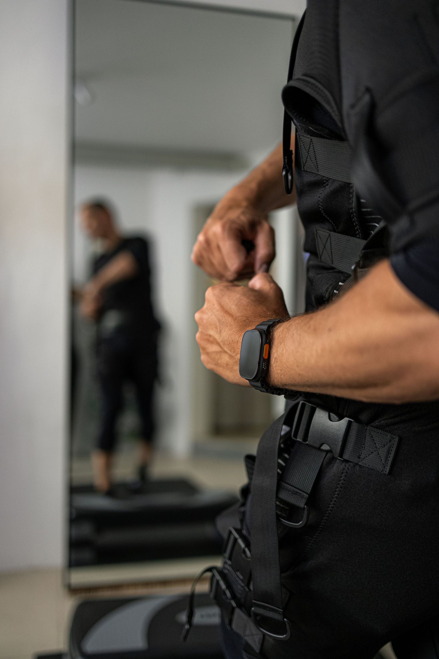 Man in black tactical vest adjusting straps, wearing a smartwatch, with mirror reflection visible in background.