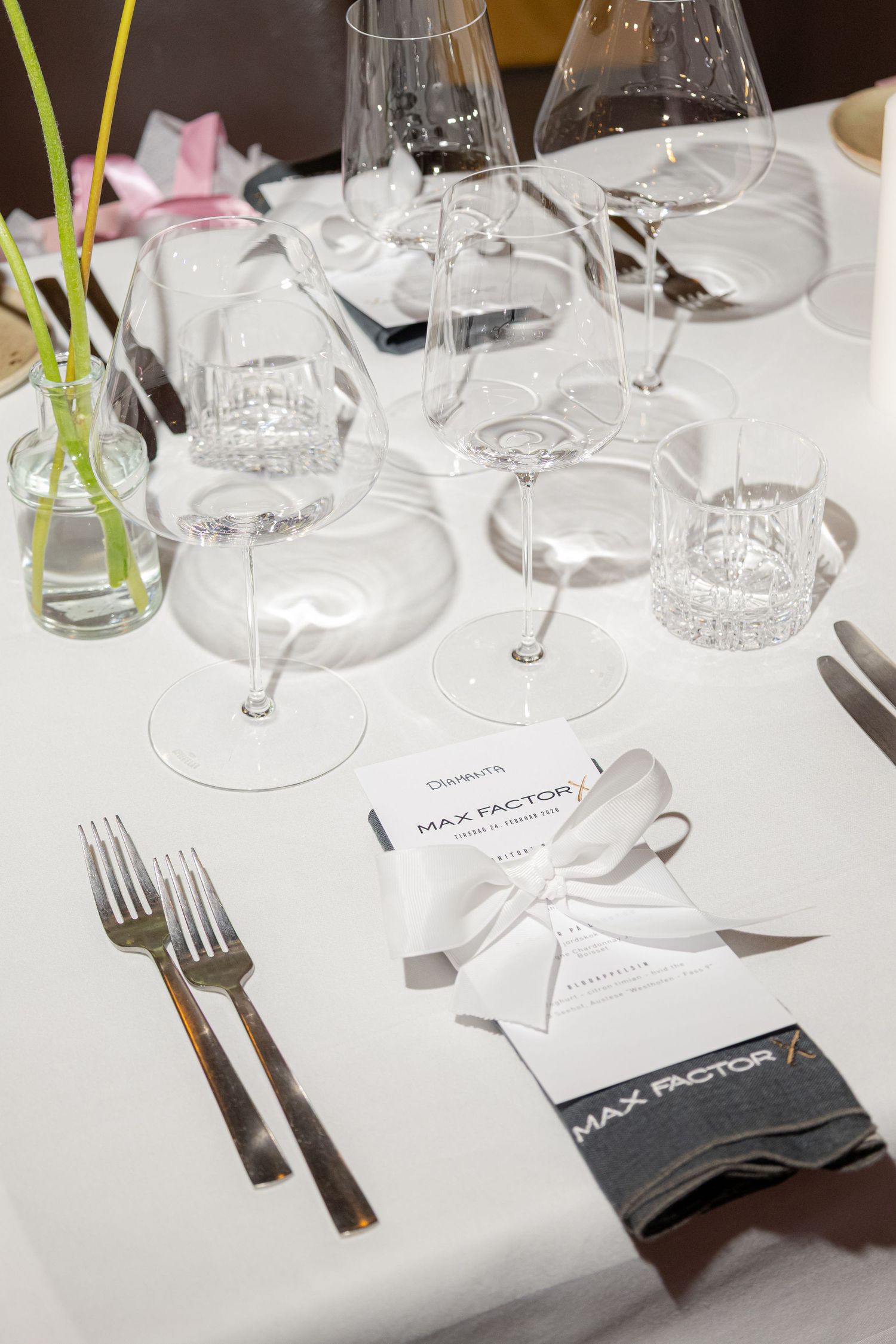 Elegant Max Factor branded table setting with crystal glasses, white linens, silverware, and decorative bow accents.