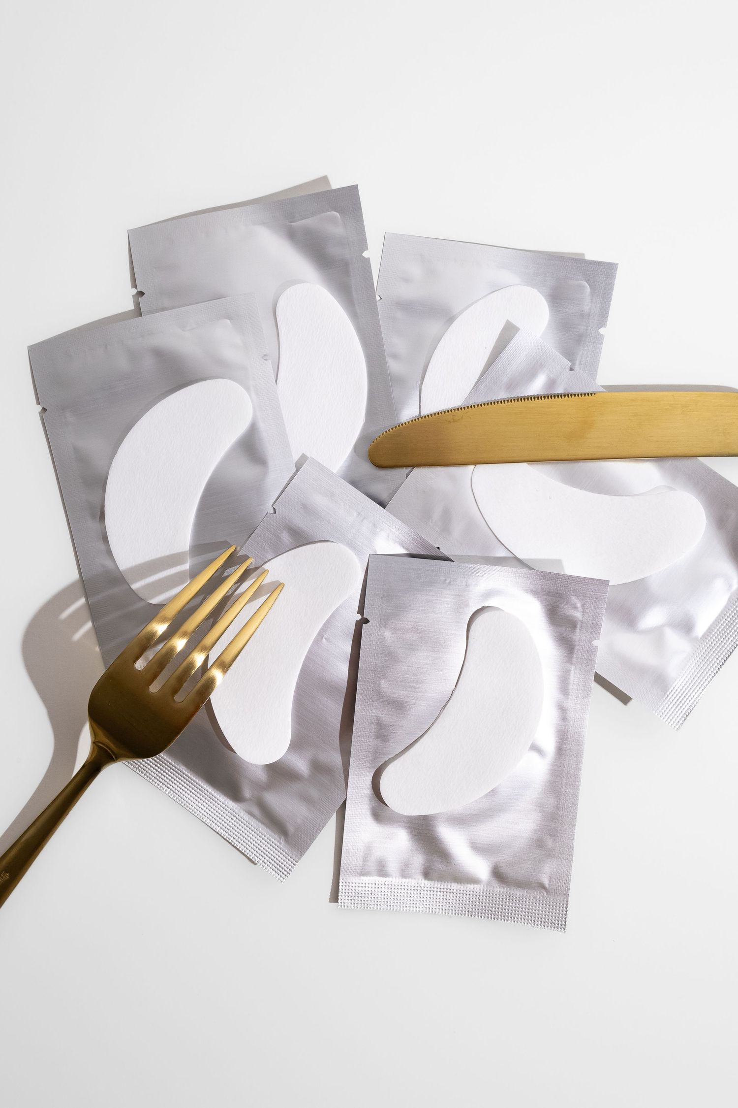 Under-eye gel patches in silver foil packets with gold fork and spatula on white background.