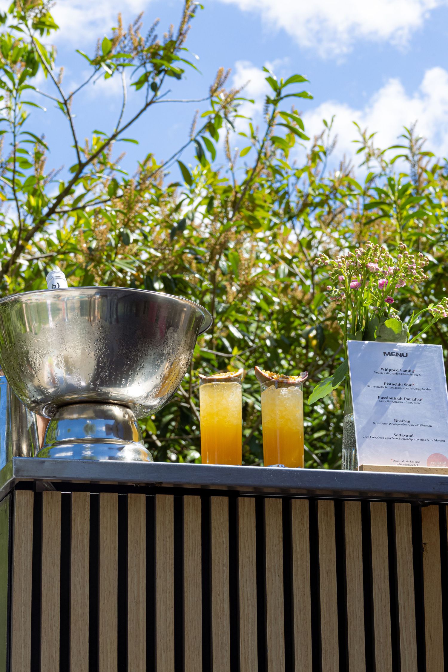 Outdoor bar setup with orange cocktails, silver ice bucket, and menu card against lush green trees.