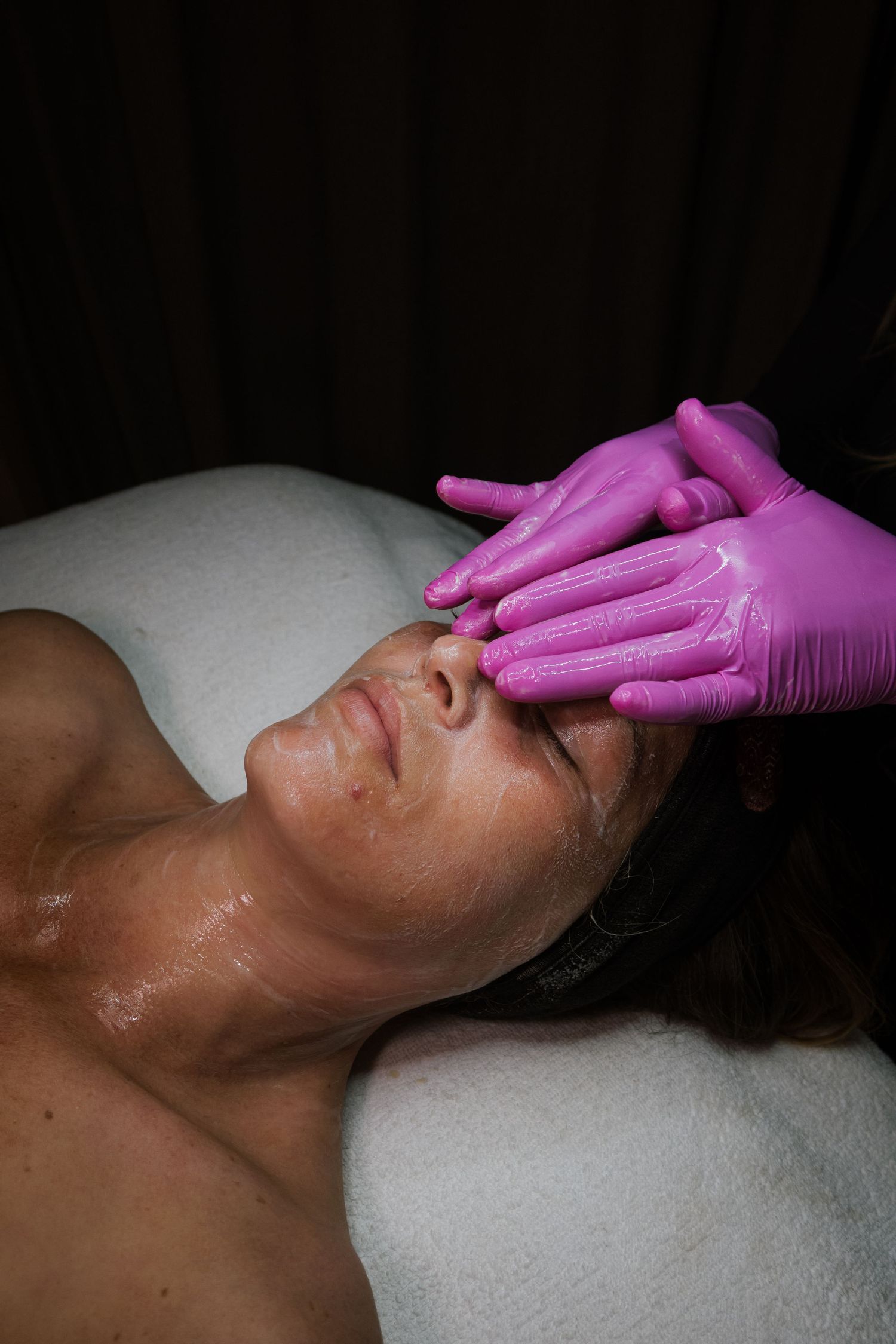 Esthetician wearing pink gloves applying facial treatment to a woman lying on a spa table.