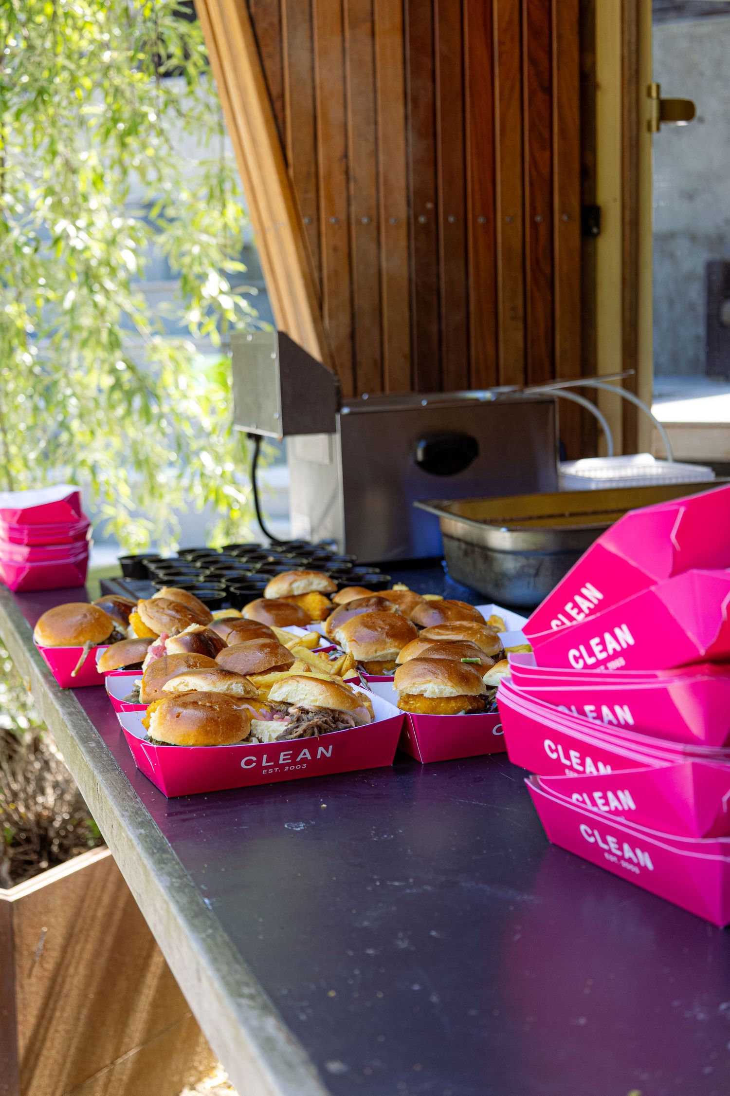Pink branded CLEAN trays of sliders on an outdoor catering table with stacked pink boxes and cooking equipment.