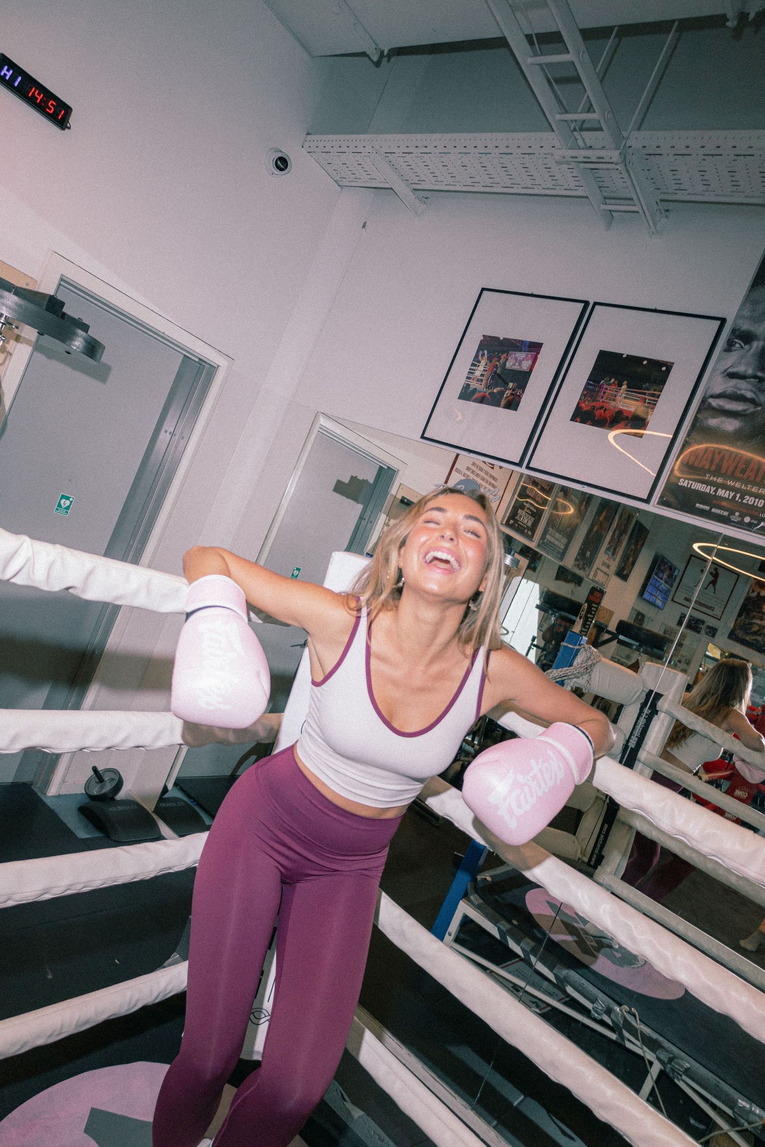 Smiling woman in pink boxing gloves and purple leggings celebrating in a boxing ring at a fitness gym.