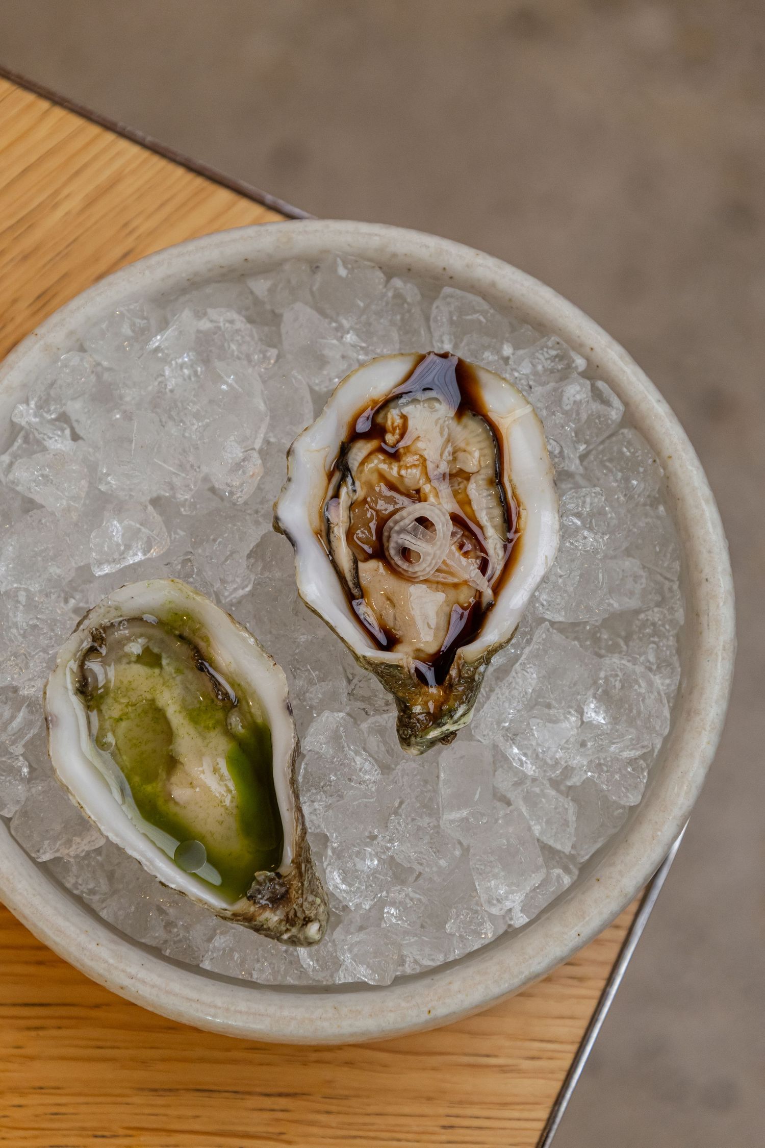 Two fresh oysters on ice in a ceramic bowl, served on a wooden board at a seafood restaurant.