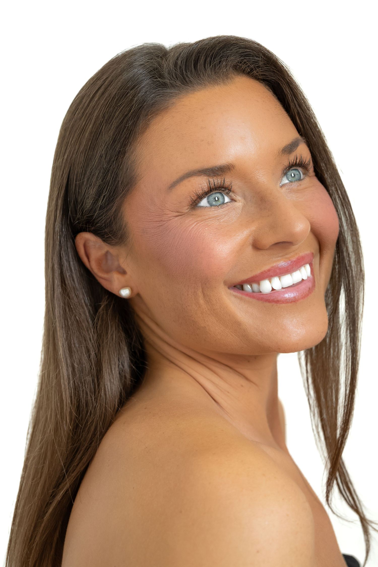 Smiling brunette woman with blue eyes and pearl earring, glowing tan skin, long straight hair, white background.