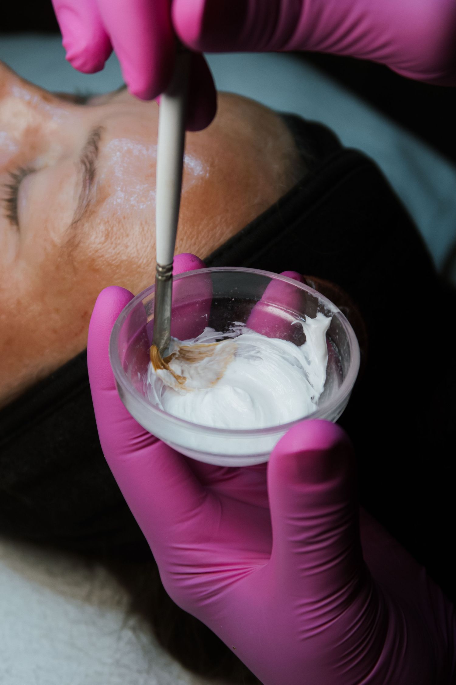 Esthetician in pink gloves applying white facial mask with brush from glass bowl to client's face.