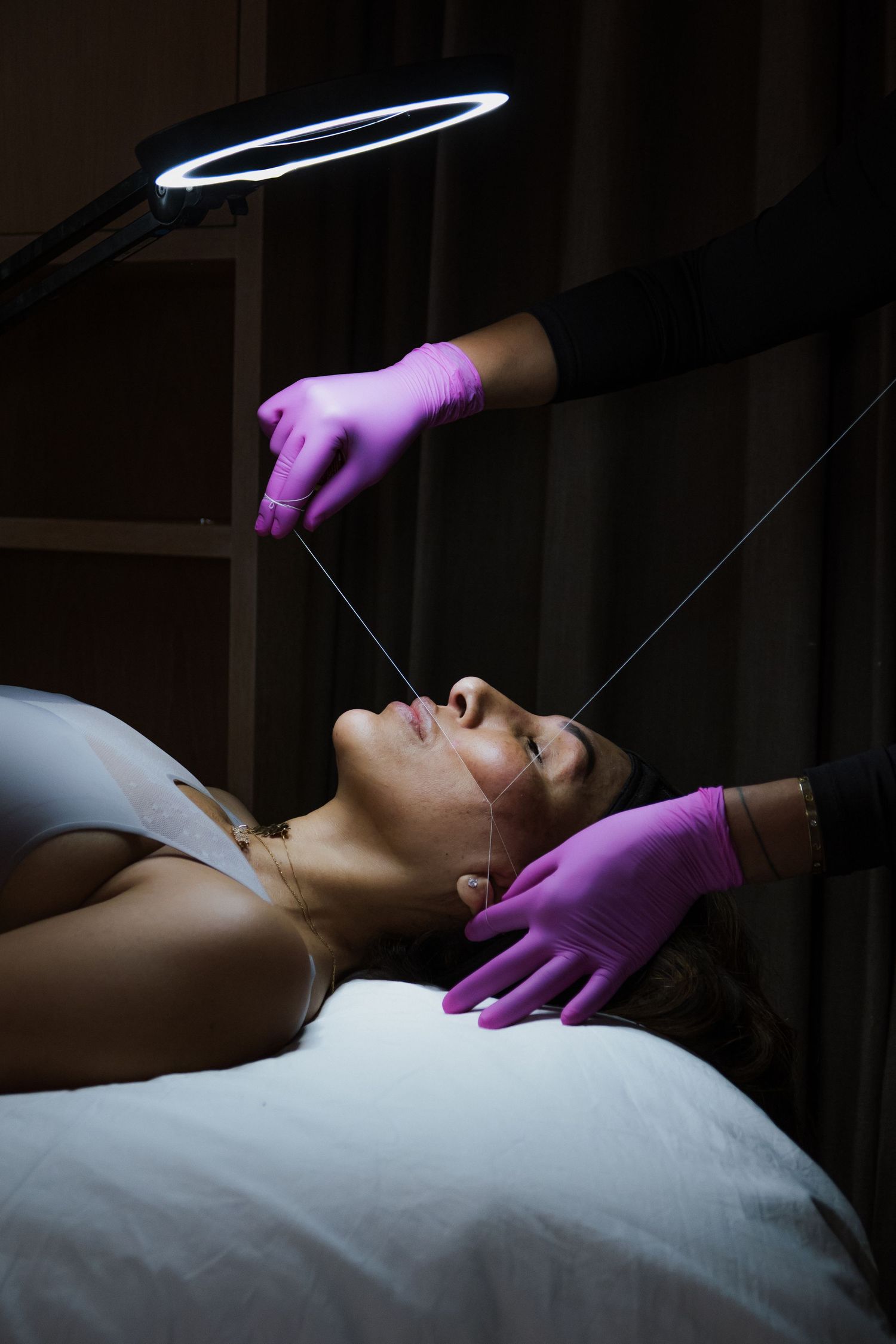 Esthetician in pink gloves performing facial threading treatment on woman lying on spa table under ring light.