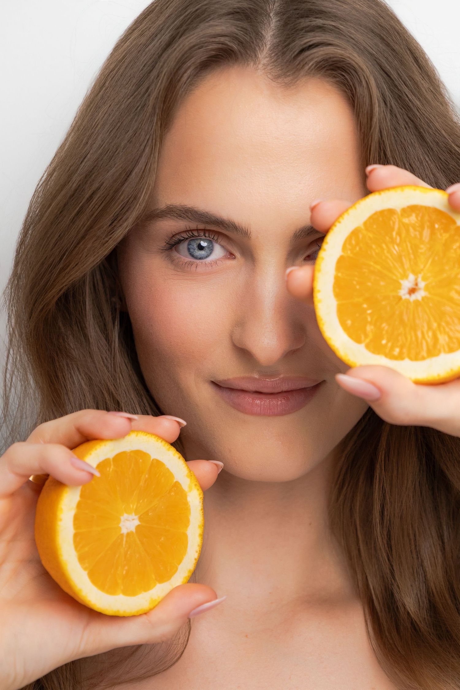 Woman holding two orange halves up to her face, showcasing fresh citrus fruit against a white background.
