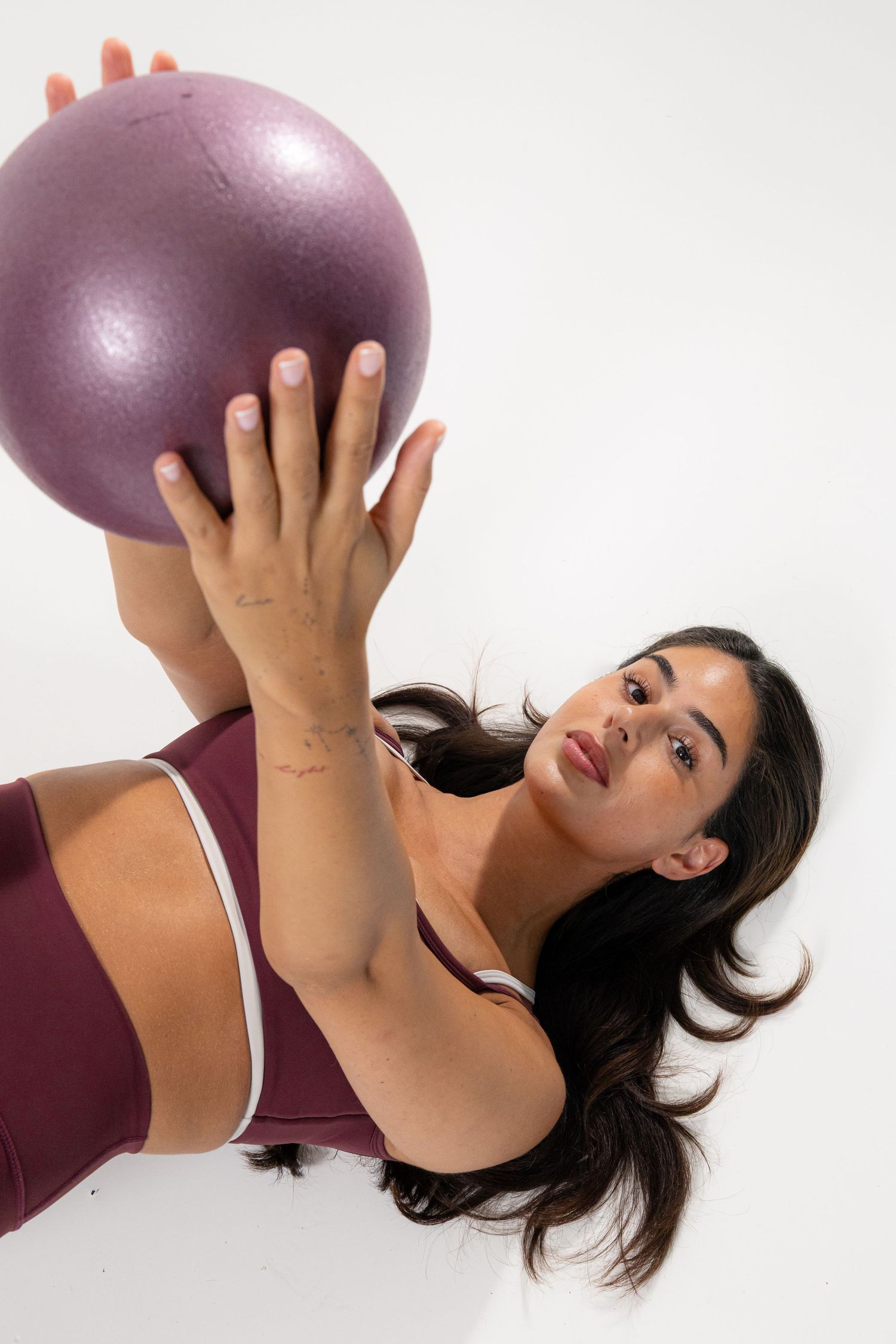 Woman in burgundy sports bra holding a shiny purple pilates ball overhead while lying on a white background.
