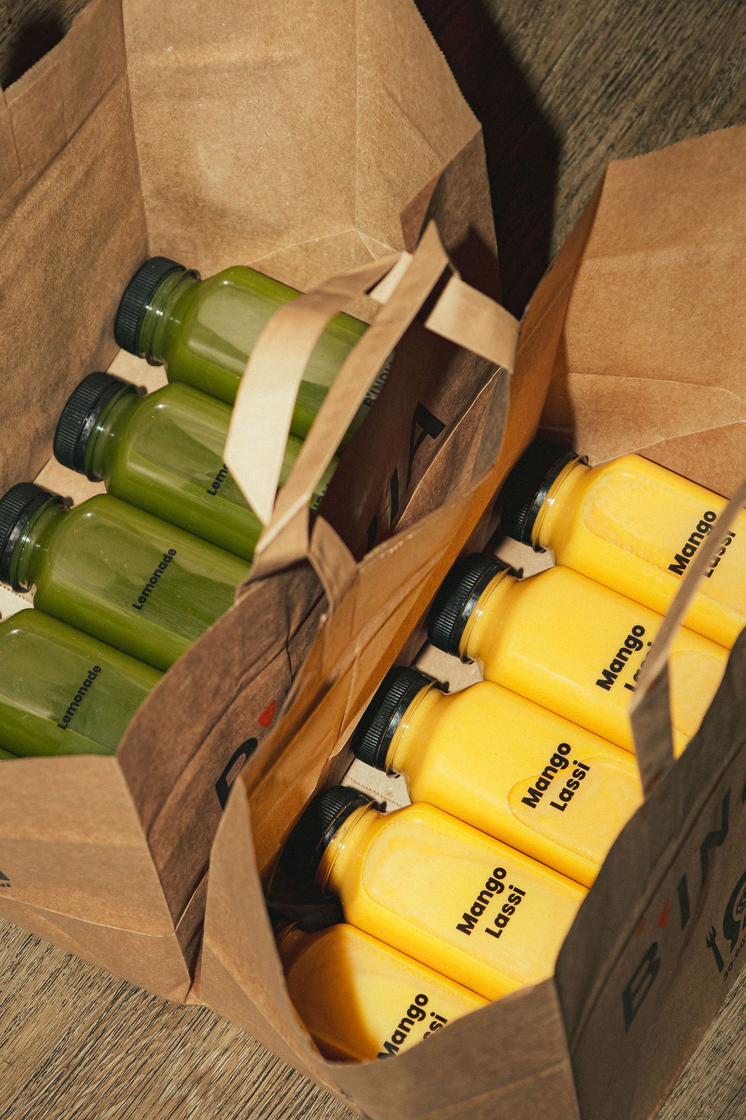 Two paper bags filled with green juice bottles and yellow Mango Lassi drinks packed for delivery.