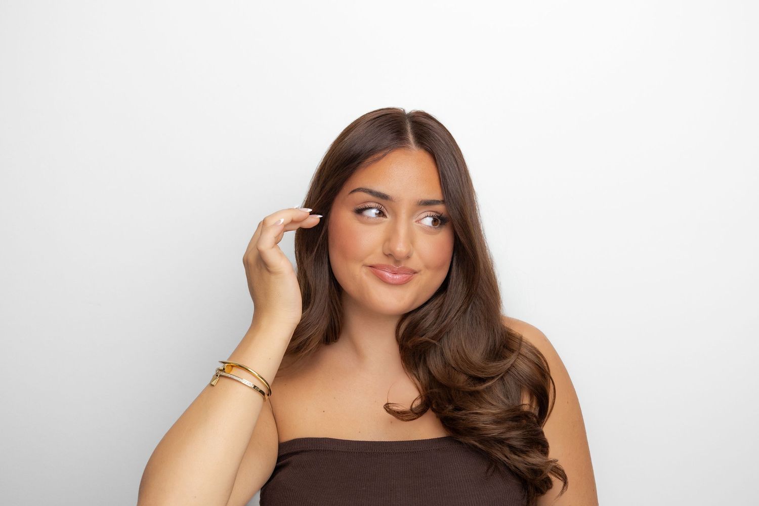 Young woman with long brown hair in brown strapless top, touching her temple thoughtfully against white background.