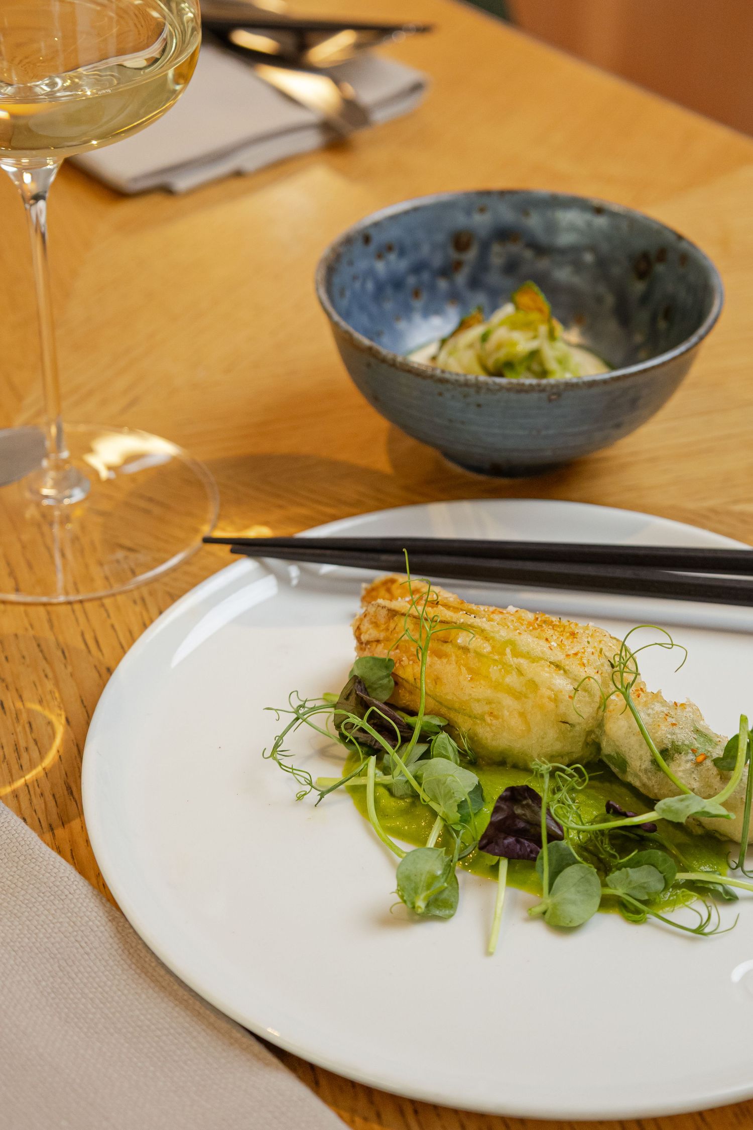 Elegant fine dining plate with seared fish, microgreens, and chopsticks, served with white wine and a ceramic bowl.