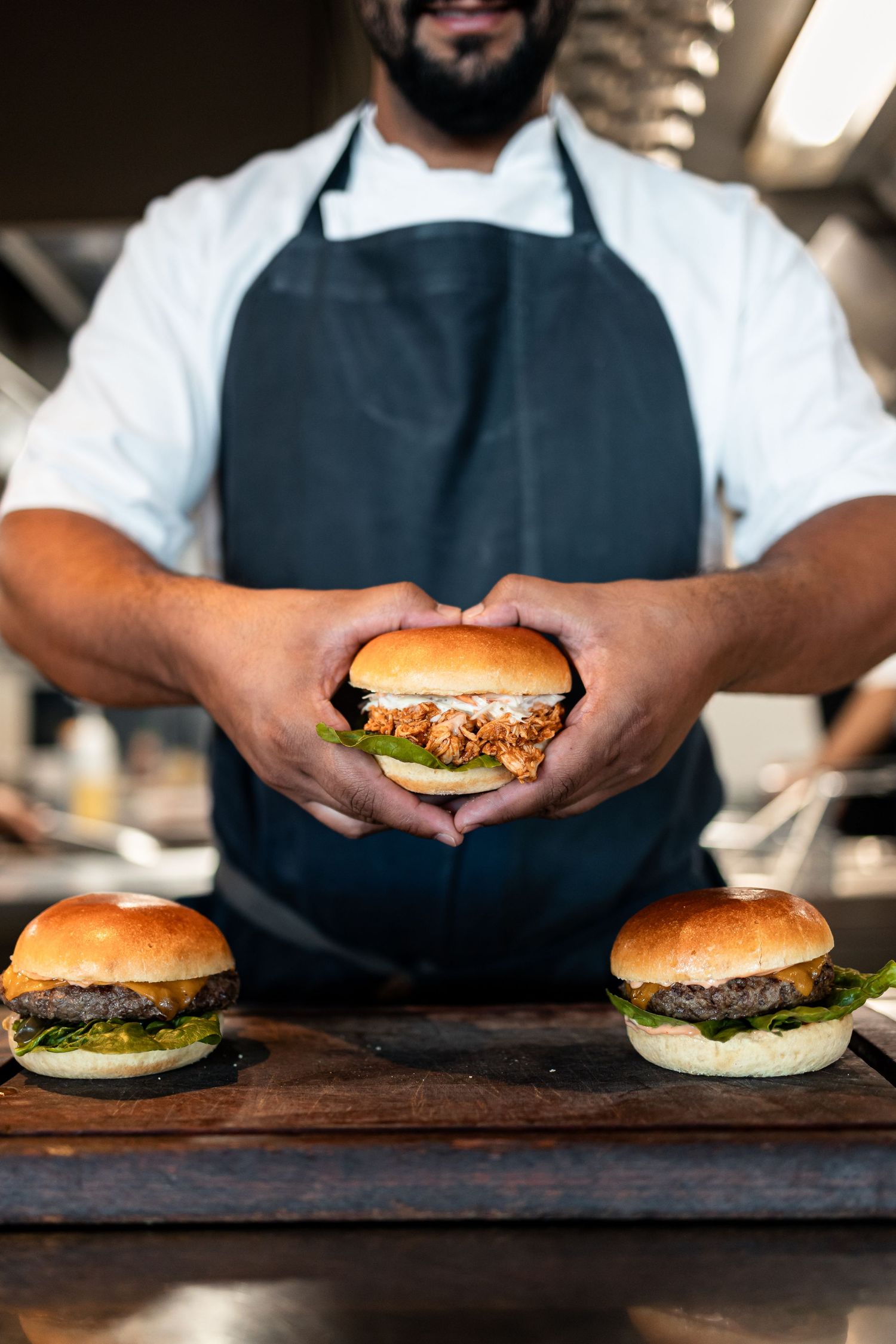 Chef in dark apron holds a pulled pork burger over a wooden board with two classic beef burgers in a restaurant kitchen.