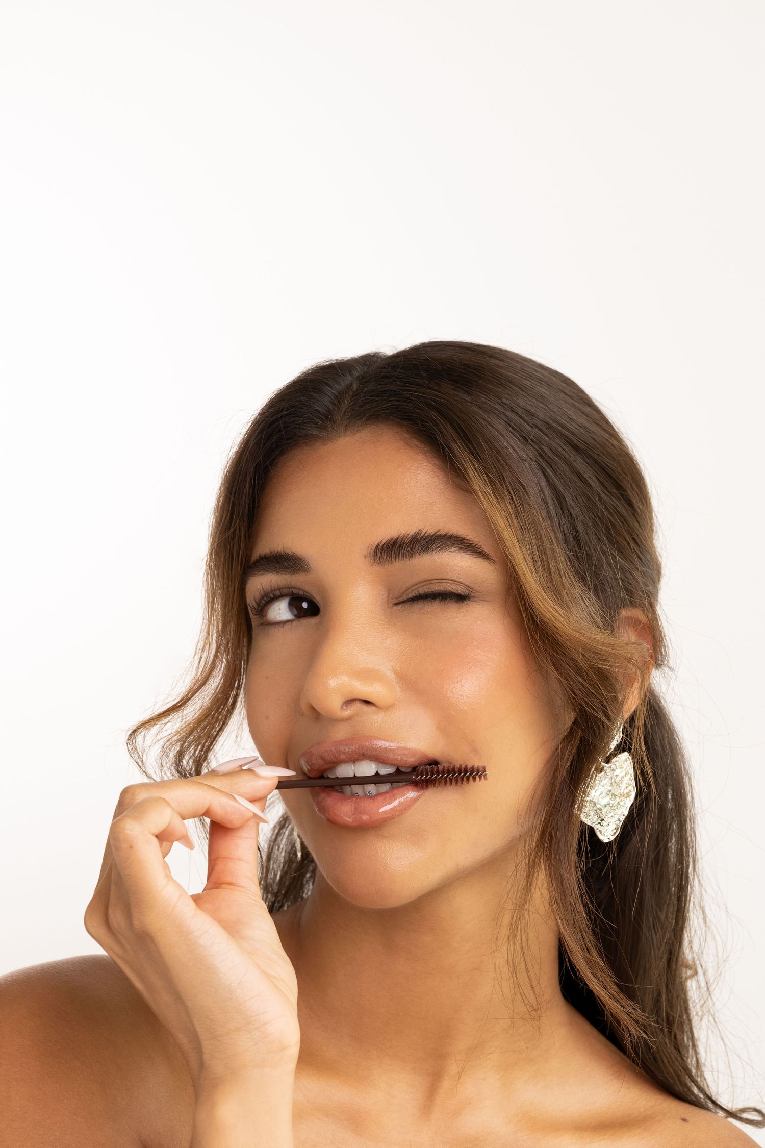 Young woman winking and holding a lash comb to her lips, wearing statement earrings against white background.