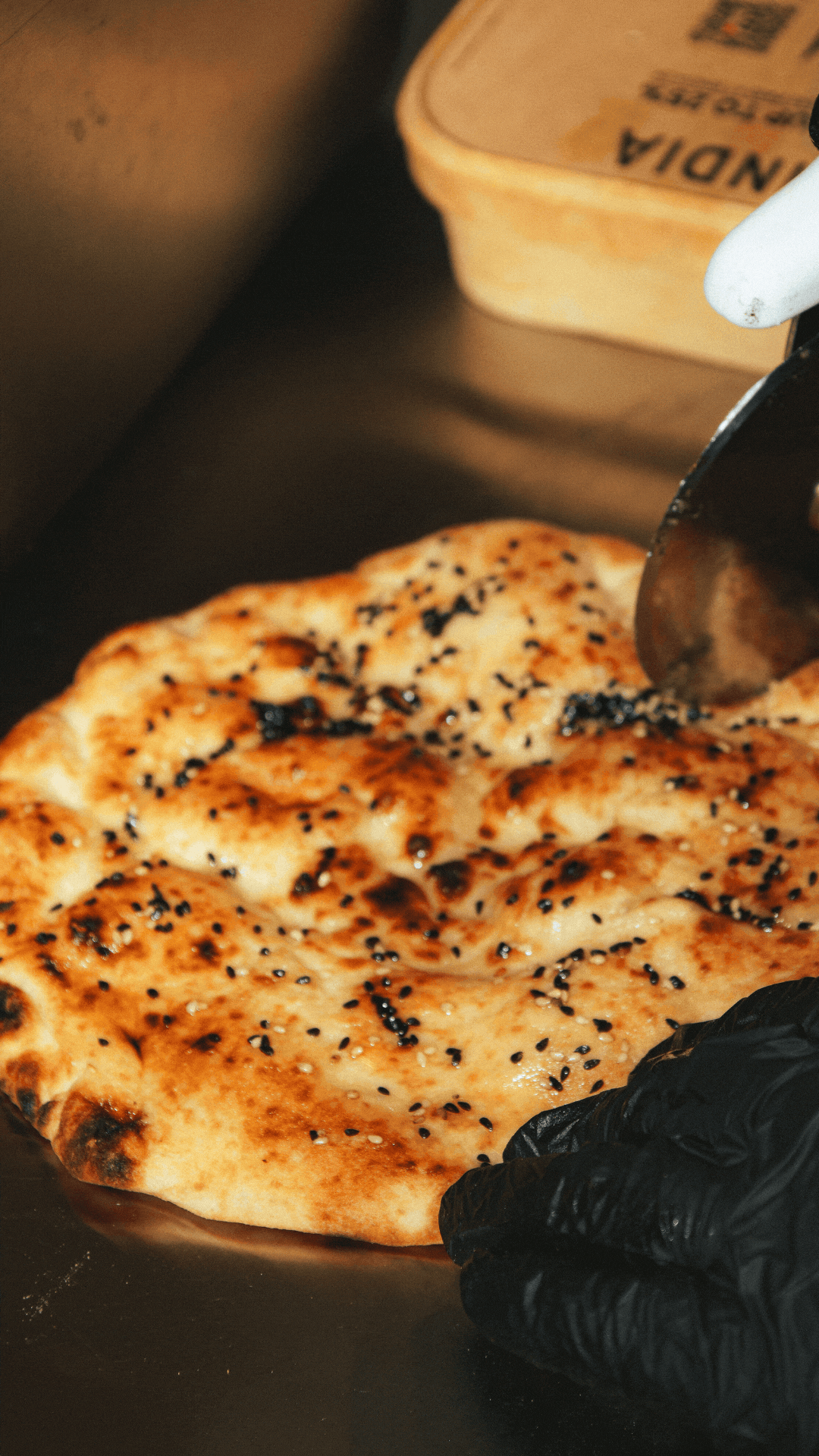 Freshly baked naan bread topped with black sesame seeds on a dark surface with gloved hands visible.