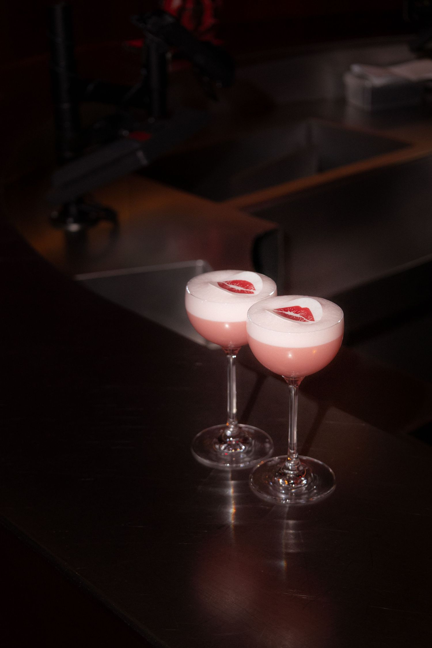 Two pink cocktails with frothy tops and red lip decorations sit in elegant stemmed glasses on a dark wooden surface.