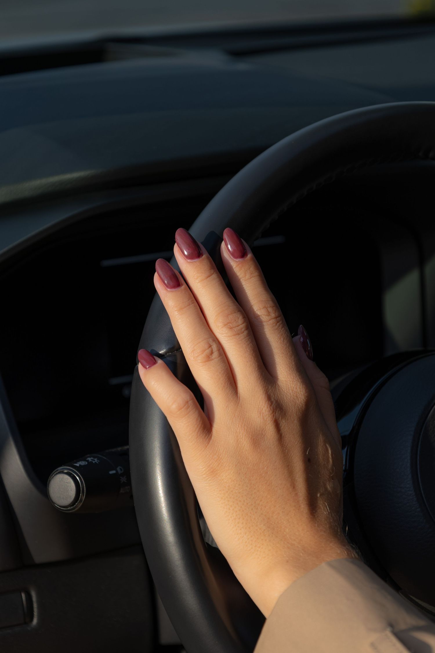 Close-up of a womans hand with dark burgundy nails resting on a black car steering wheel.