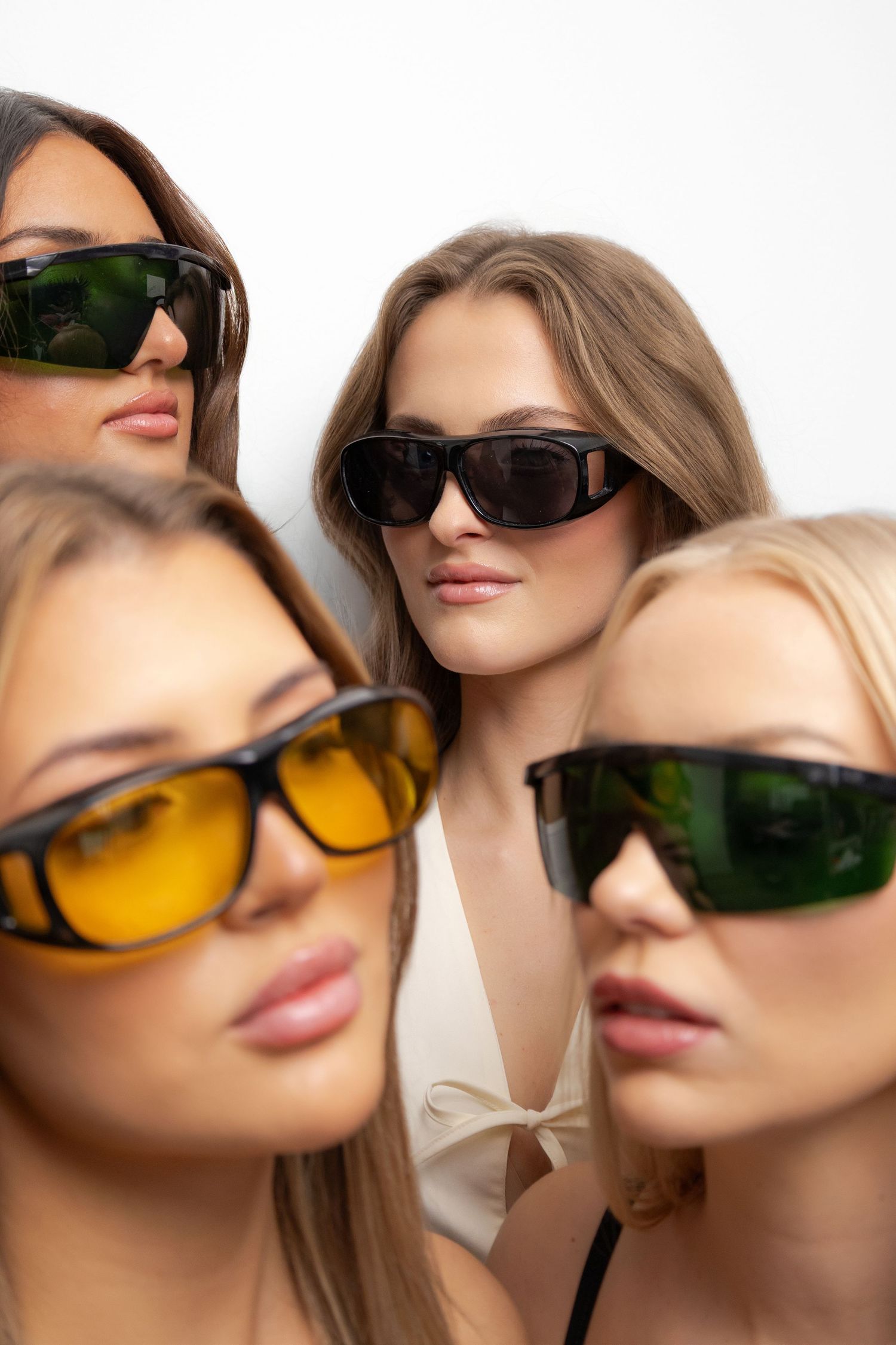 Four women wearing stylish sunglasses with colored lenses posing together against a white background.