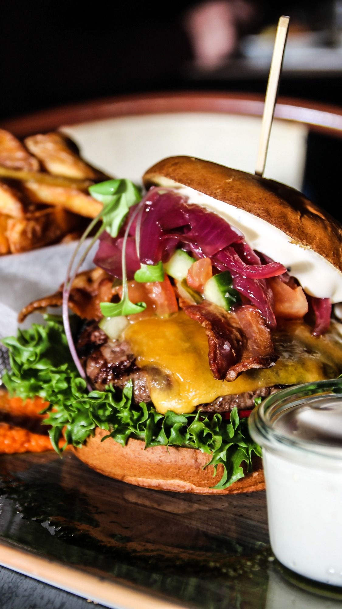 Gourmet burger with melted cheese, red onions and greens served on dark slate with white sauce.