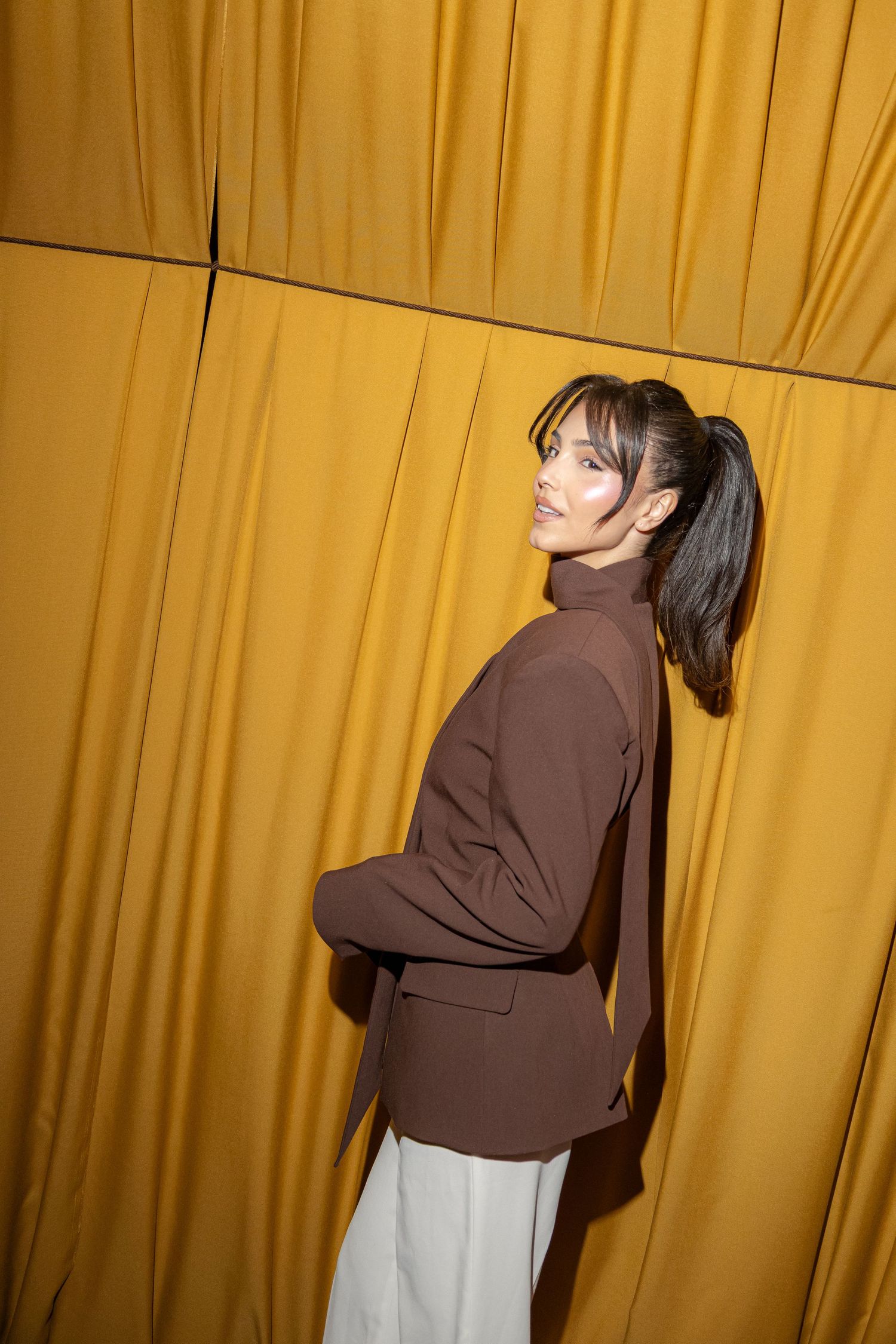 Woman in brown blazer and white pants poses against golden yellow curtain backdrop.