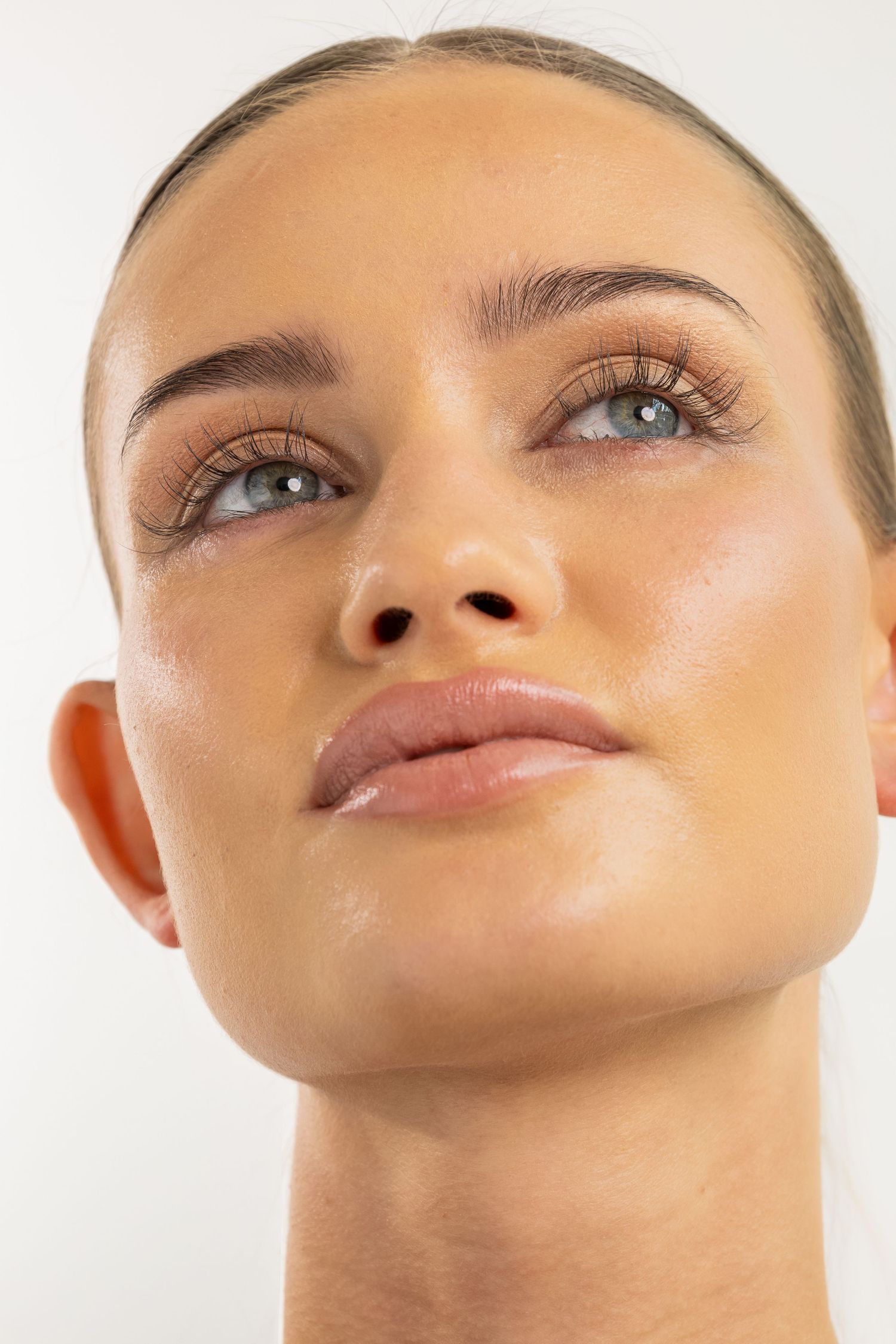 Close-up portrait of a young woman with glowing skin, subtle makeup, and long lashes gazing upward.