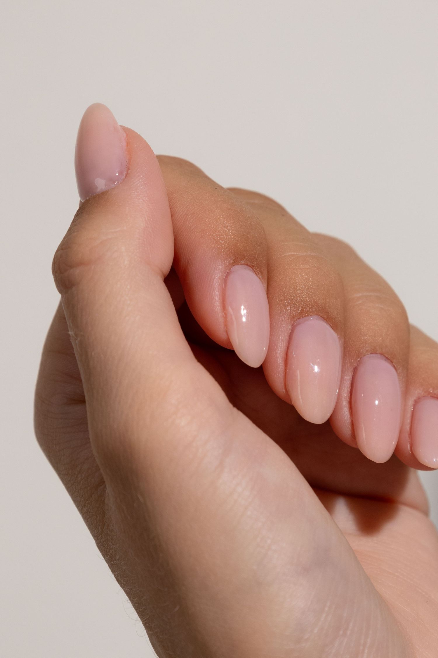 Close-up of manicured hands with glossy pale pink oval nails against a neutral background.