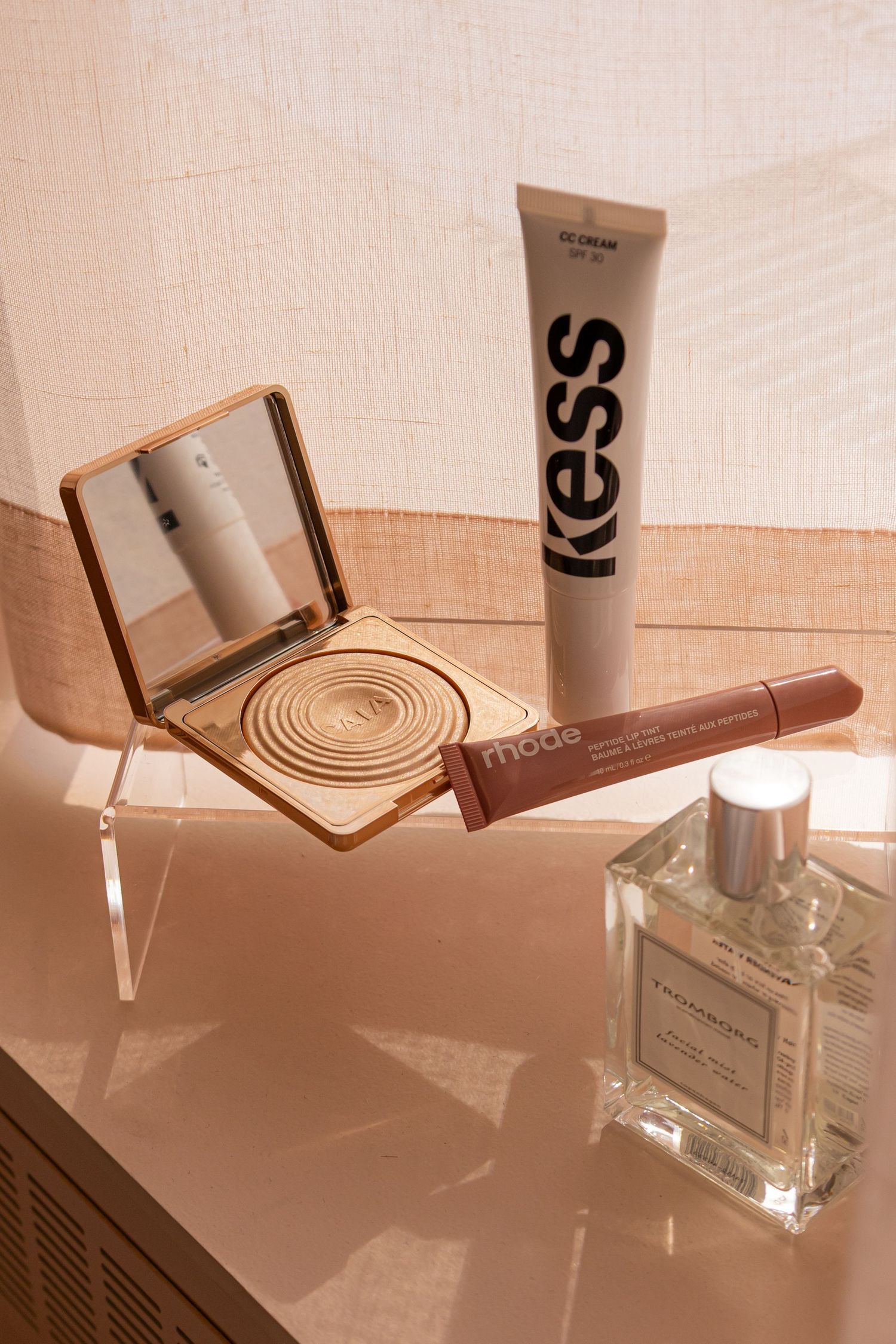 Flat lay of Kess BB cream, Mode lip liner, gold compact mirror, and glass perfume bottle on a pink surface.
