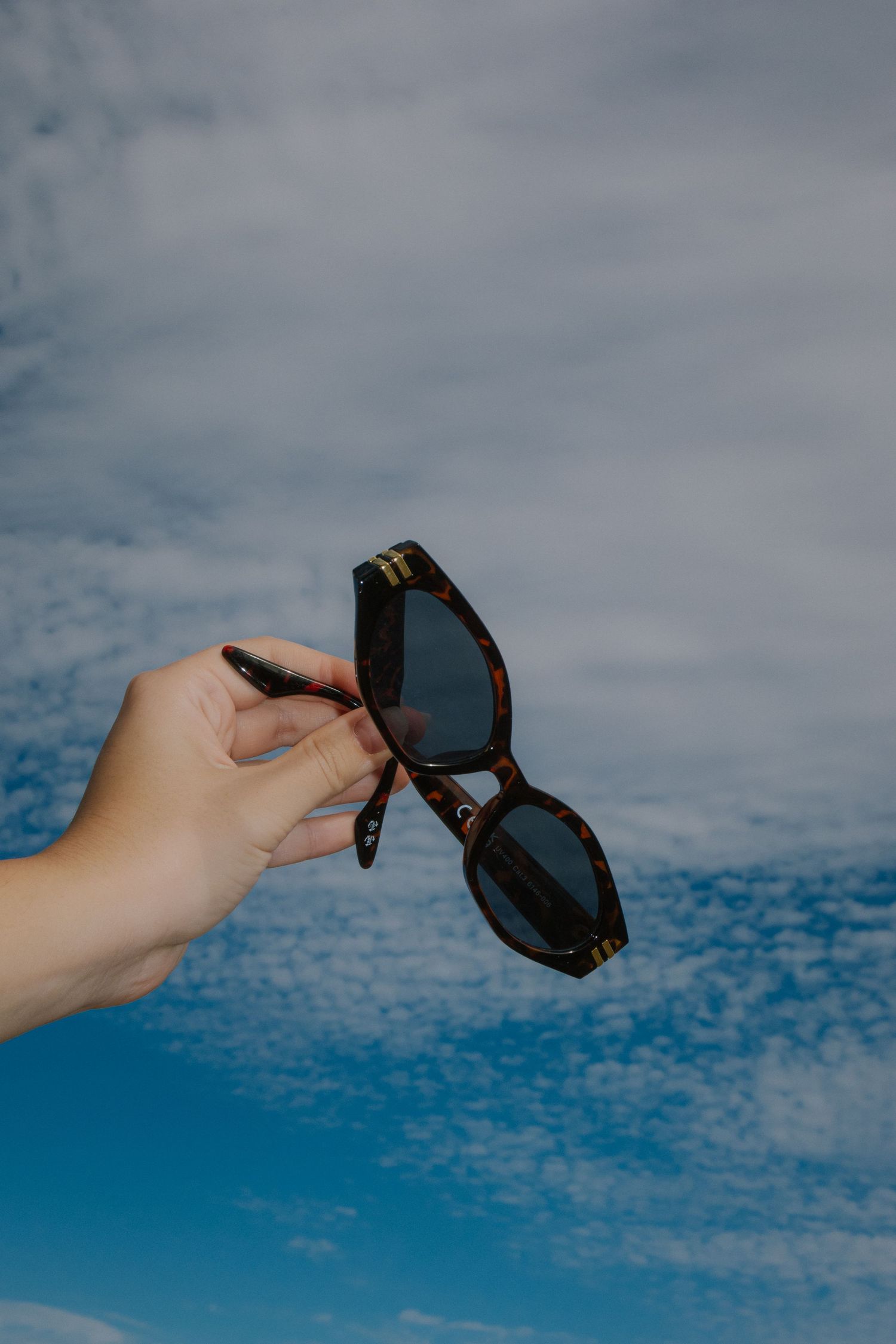 Hand holding sunglasses against blue sky with scattered clouds.