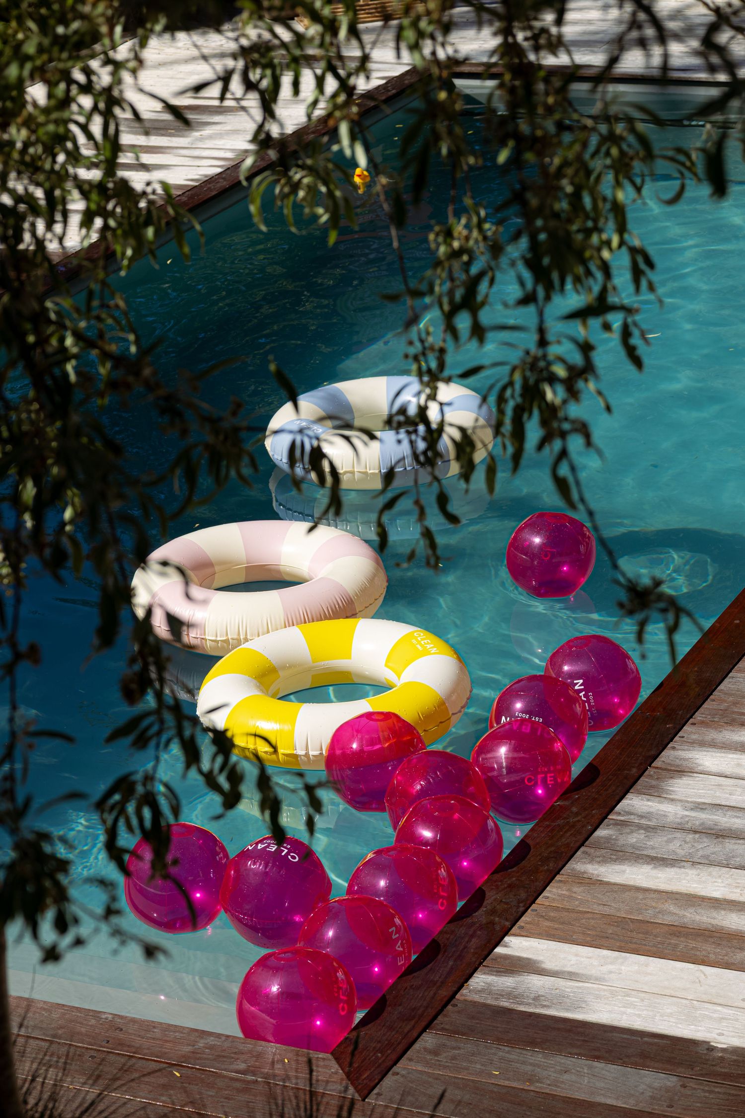 Colorful inflatable pool rings and pink water balloons floating near a wooden deck in a sunny blue swimming pool.