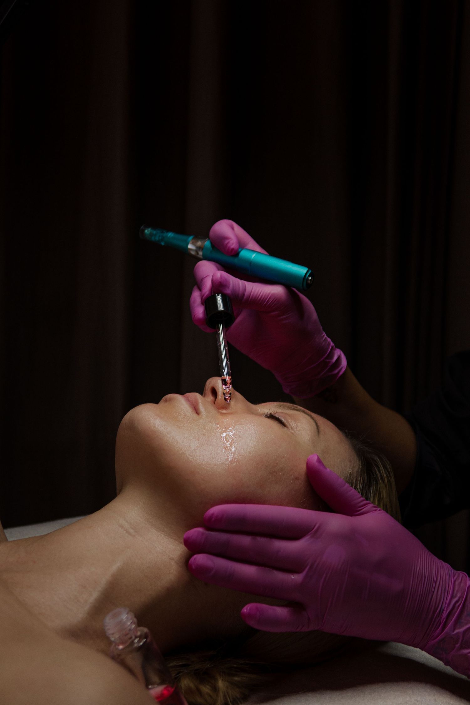 Esthetician in pink gloves performing microneedling treatment on woman's face with dermapen device in spa.