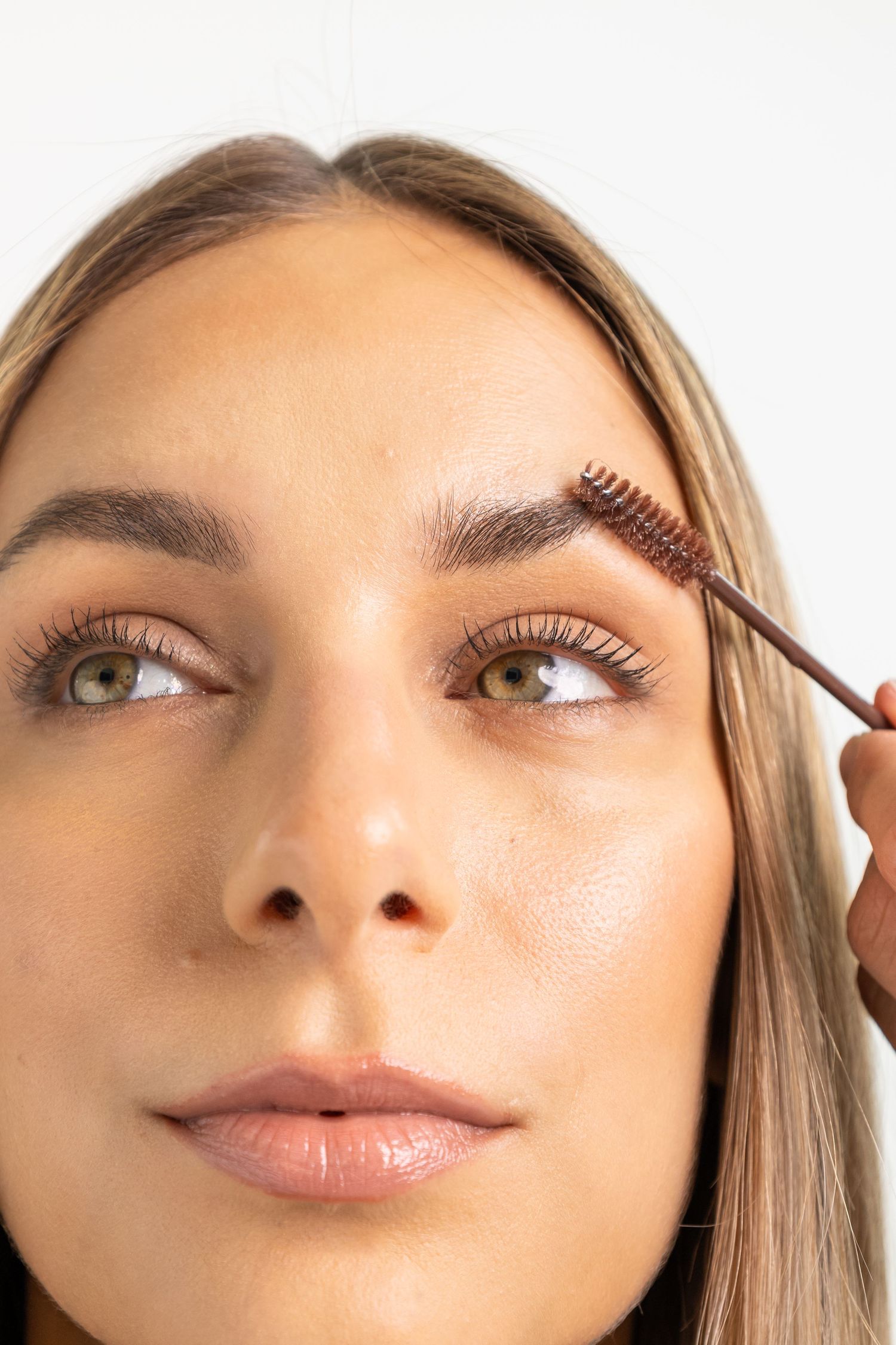 Close-up of a woman grooming her eyebrows with a spoolie brush, showcasing natural makeup and glowing skin.