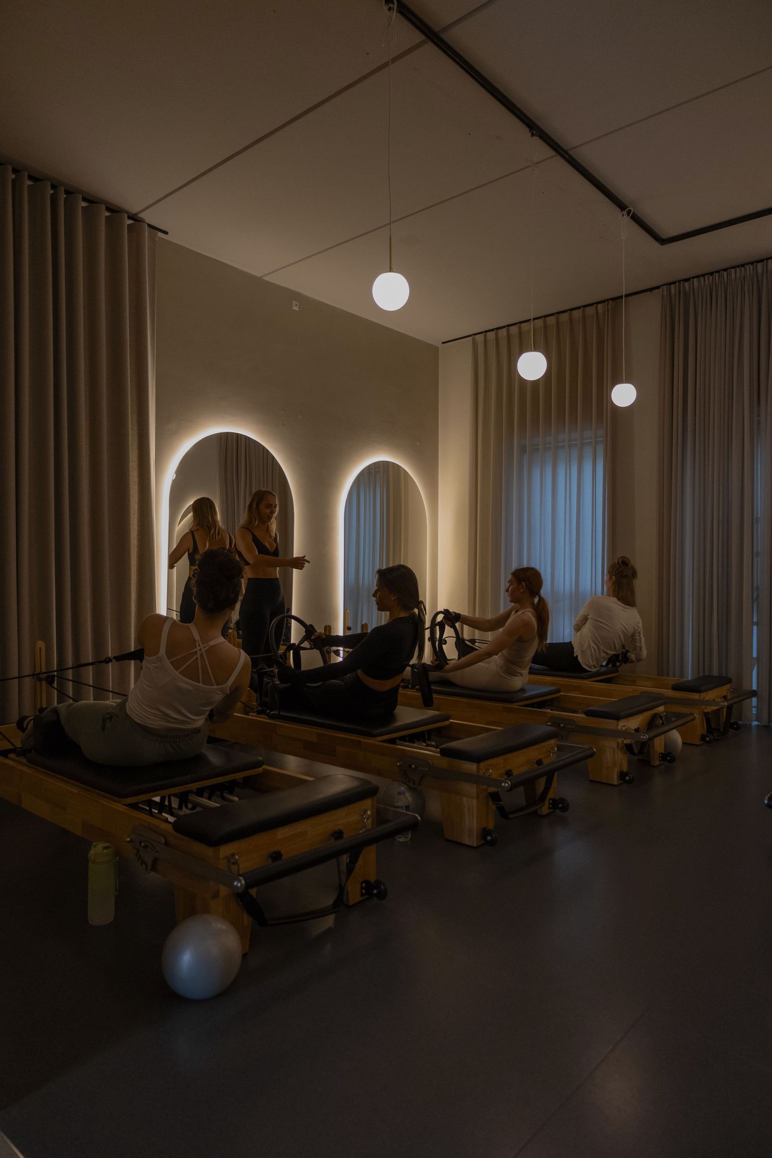 People practicing Pilates on reformer machines in a dimly lit studio with arched mirrors and pendant lights.