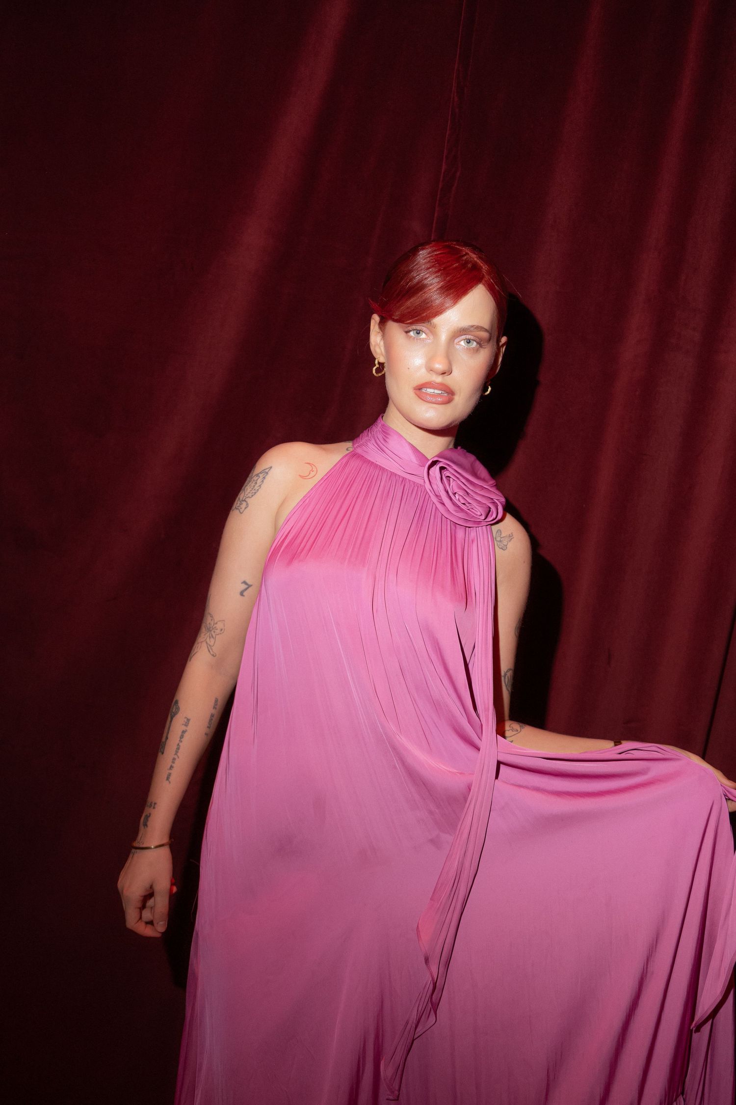 A model with red hair poses in a flowing pink halter gown with rose detail against a deep red velvet backdrop.