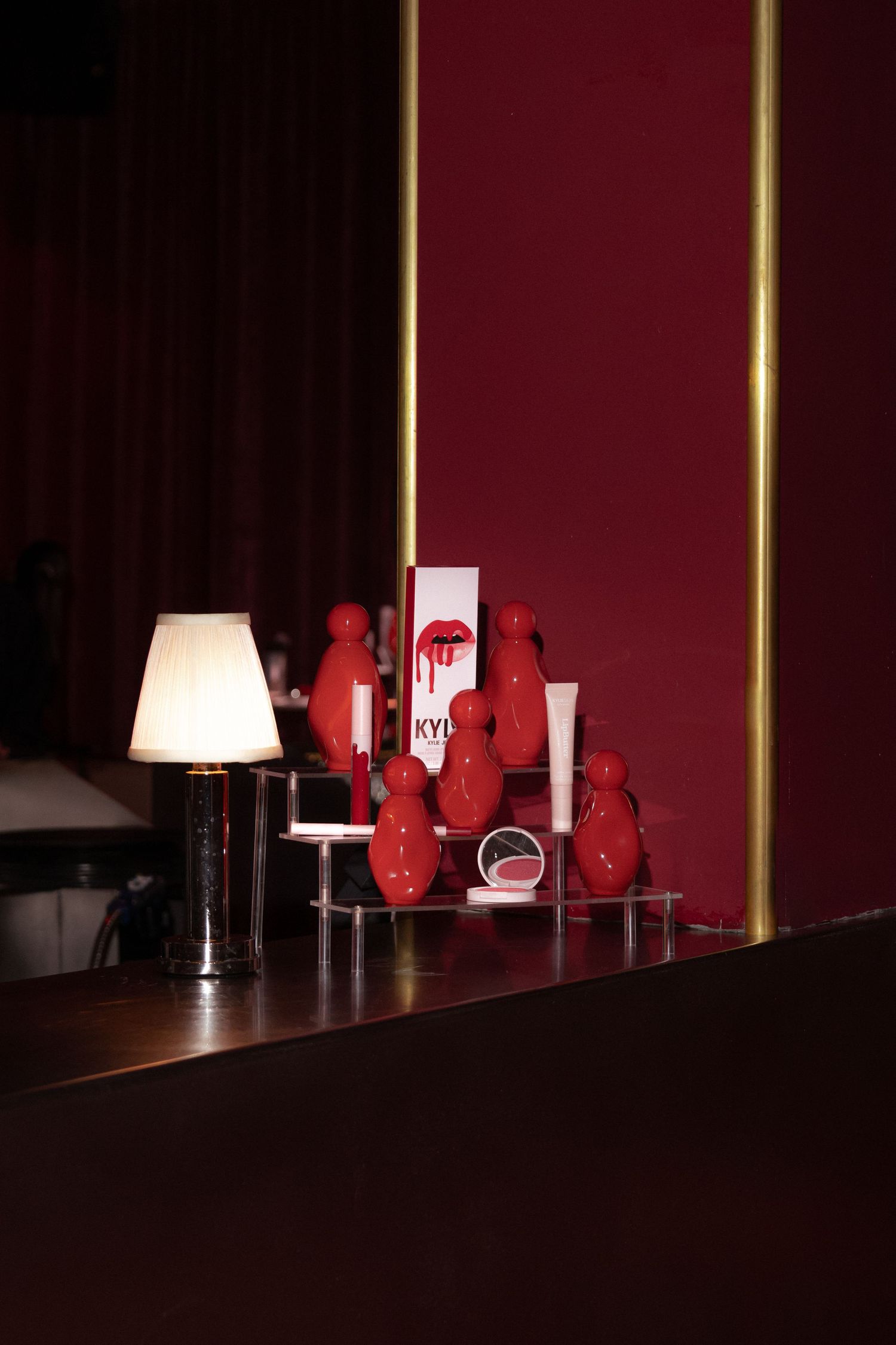 Kylie Cosmetics red lip kit display with decorative red figurines on a shelf beside a small lamp in a dark red room.